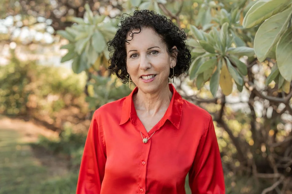 A woman with curly dark hair wearing a red blouse and earrings, posing outdoors in front of green foliage.