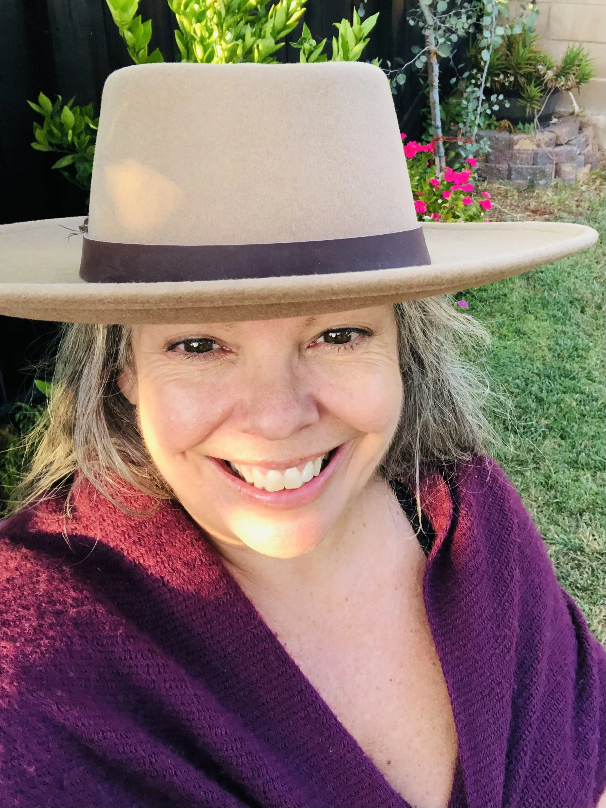 A woman smiling outdoors wearing a large beige wide-brimmed hat with a dark band, a purple sweater, with garden plants and flowers in the background.