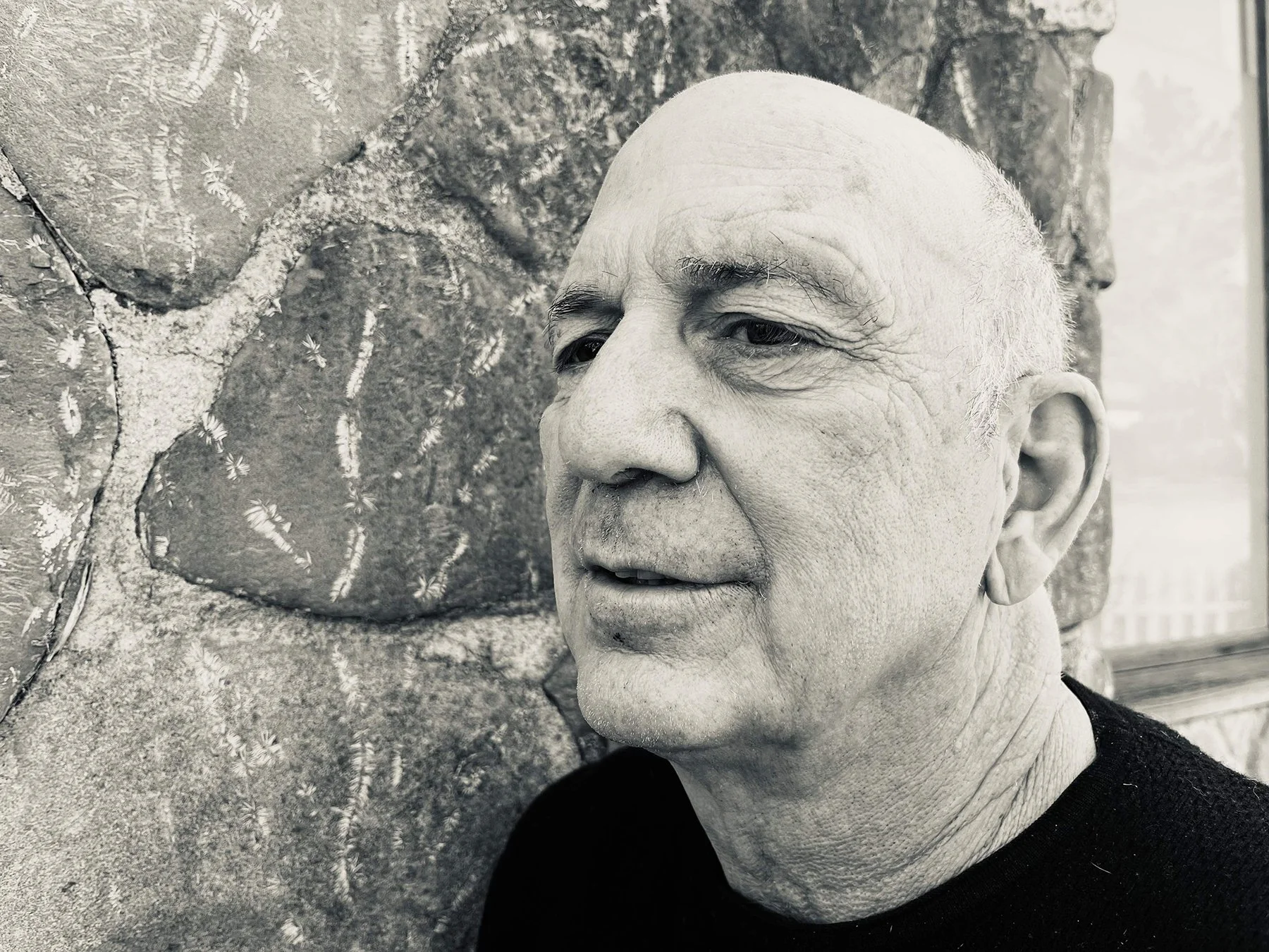 A close-up of a man with a bald head gazing to the right, standing in front of a stone wall with painted designs.