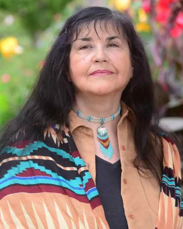 Portrait of a woman with long black hair, wearing a colorful patterned shawl, beige shirt, and turquoise necklace, outdoors with a blurred garden background.