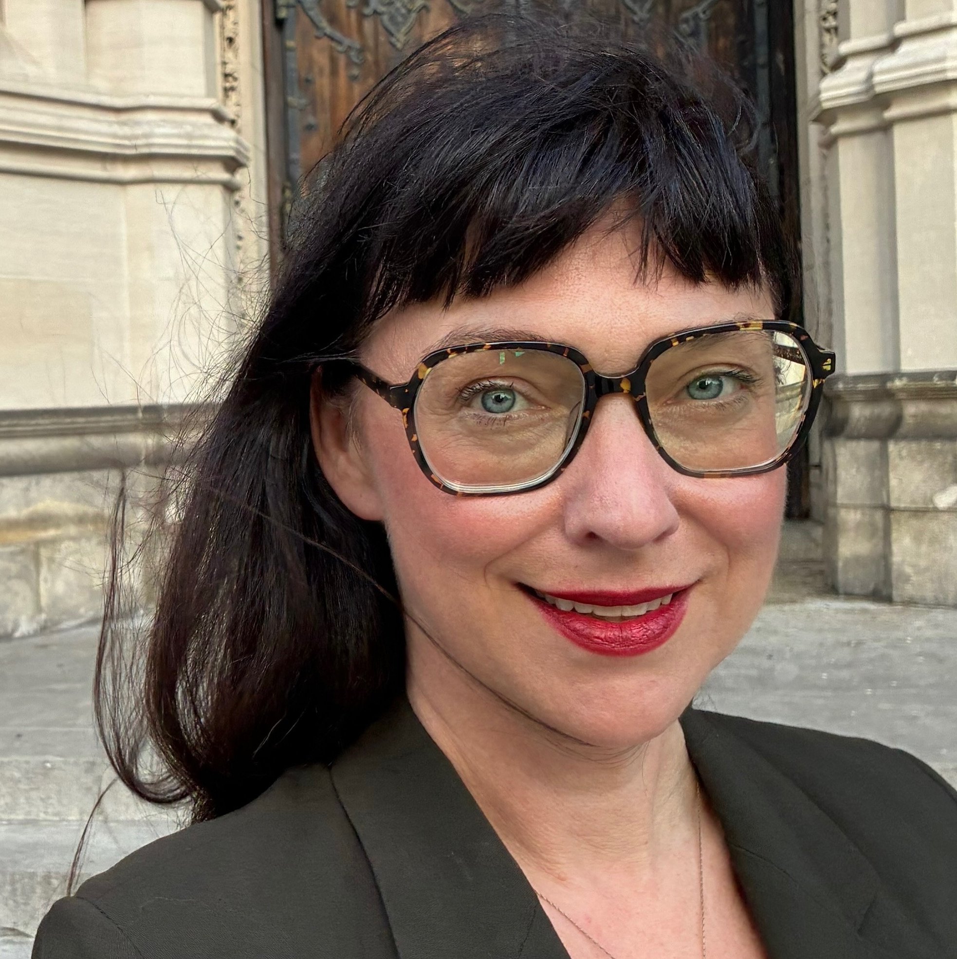 Close-up of a woman with dark hair, wearing large tortoiseshell glasses, red lipstick, and a black blazer, standing outdoors in front of a historic stone building with steps.