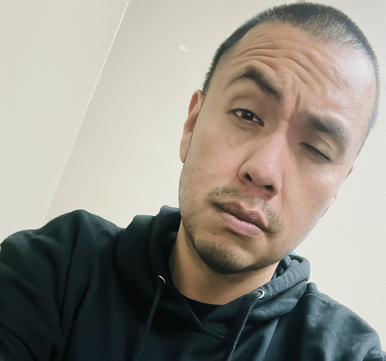 A man with short hair, facial hair, and a serious expression wearing a black hoodie, taking a selfie against a plain wall.