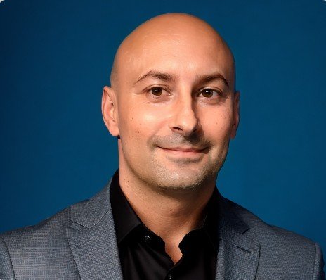 A man in a dark gray suit and black shirt posing against a blue background.