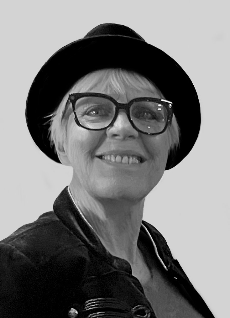 A woman with short blonde hair wearing large glasses, a black hat, and a leather jacket, smiling at the camera in black and white.