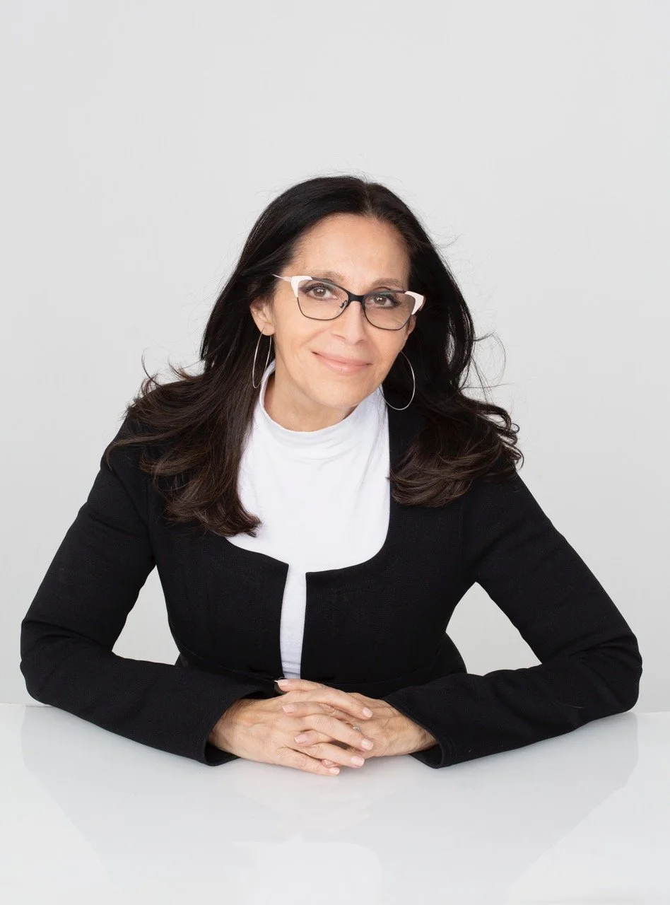 A woman with dark hair, glasses, and hoop earrings, sitting at a white table against a plain light background, smiling slightly.