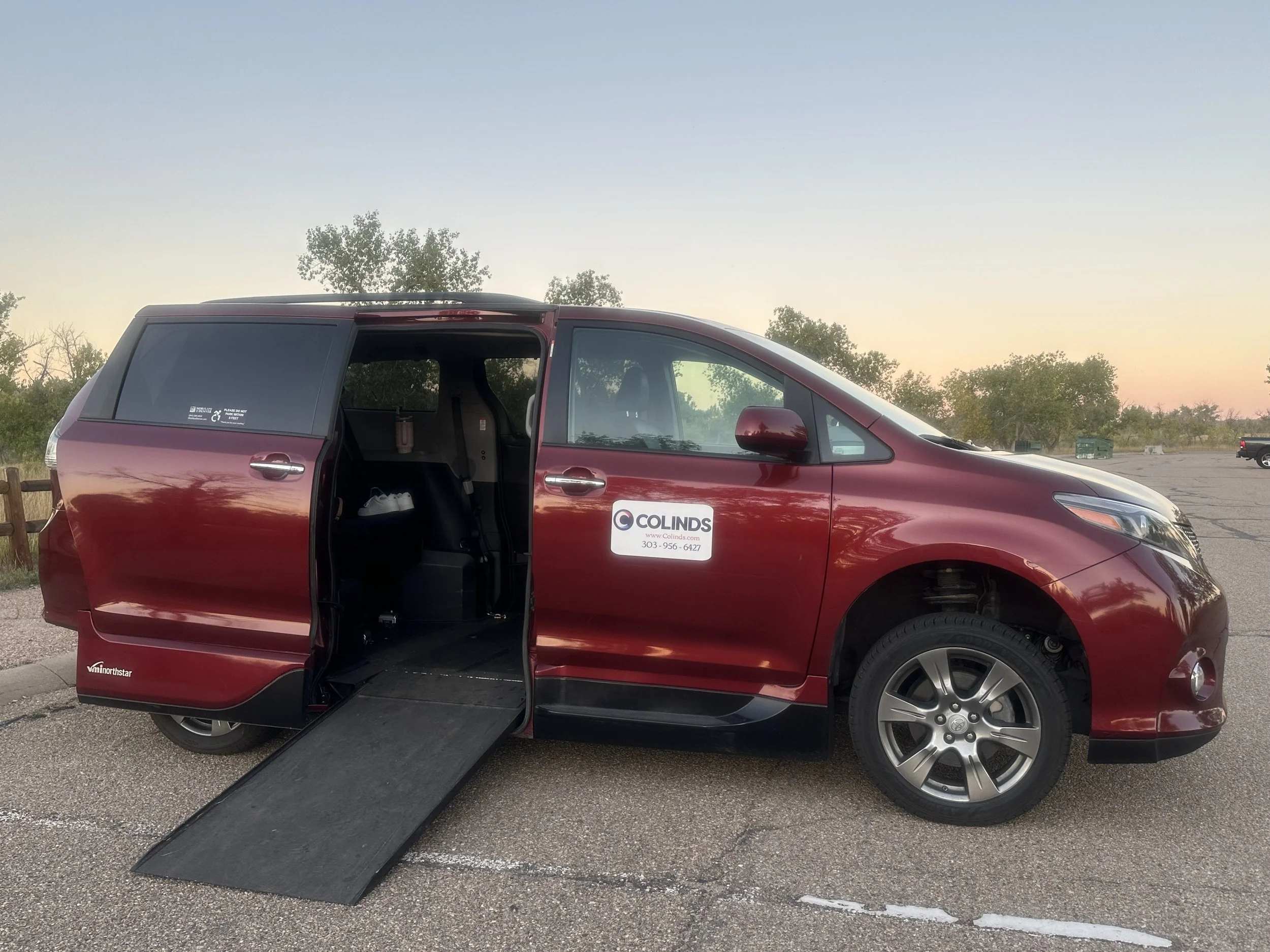 Red minivan with a wheelchair ramp deployed, parked in an open lot at sunset, with a logo for Colinds on the door.