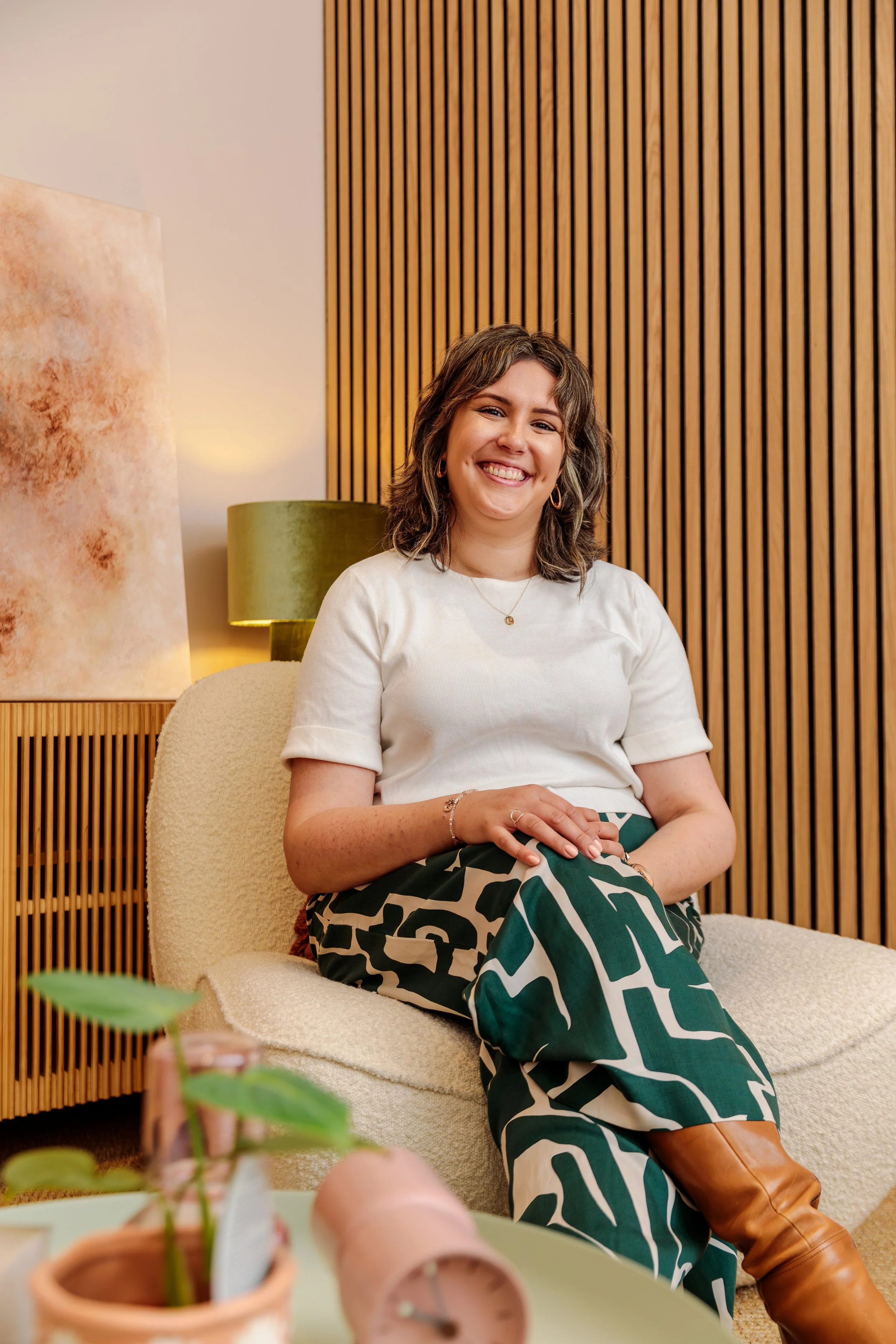 A woman sitting comfortably on a beige armchair, smiling, in a room with wooden slat wall decor and a green lampshade behind her.