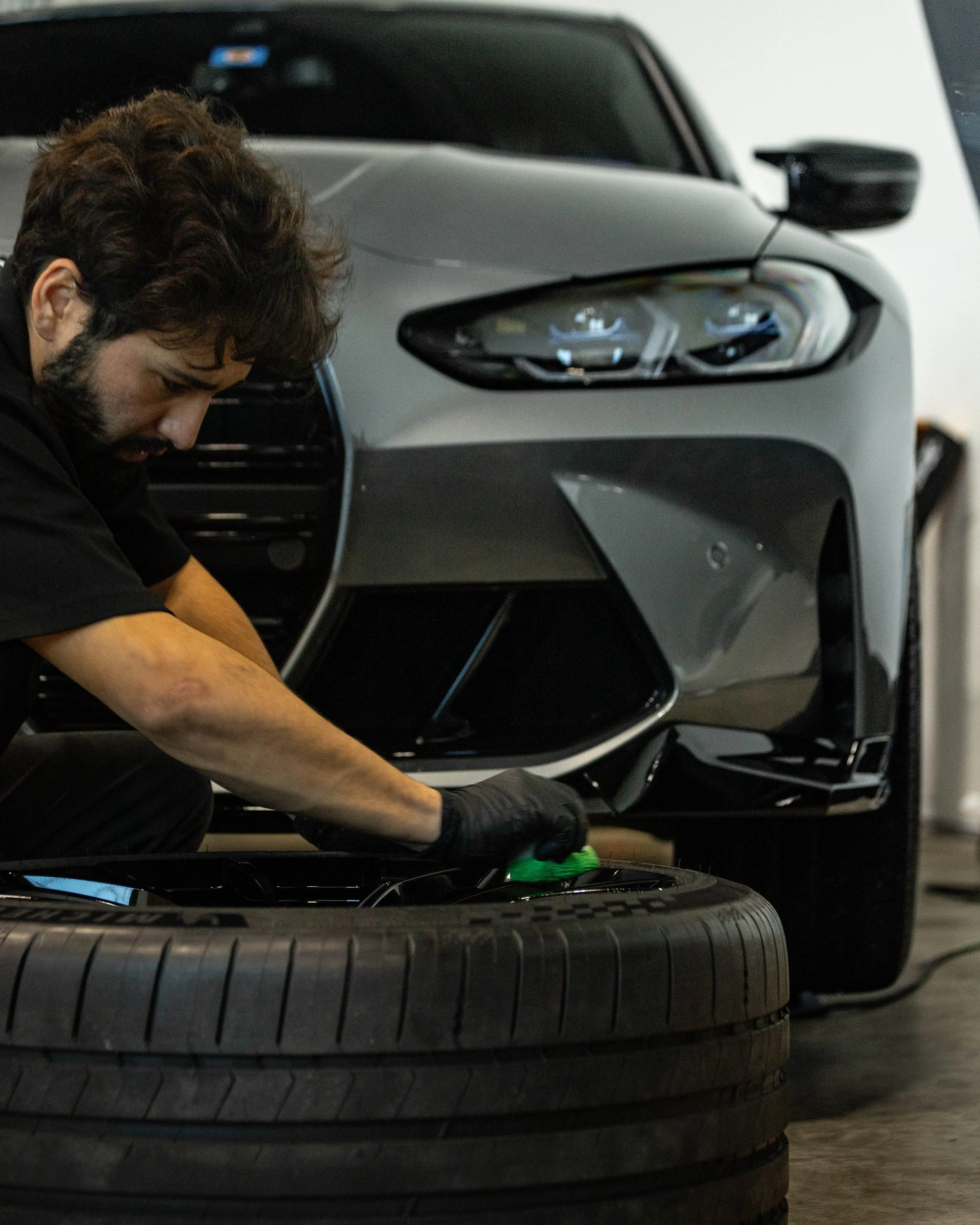A mechanic in black gloves working on a car wheel in a garage, with a gray BMW M5 elevated in the background. The wheels are recieving a ceramic coating layer for protection and a refreshed or improved gloss finish.
