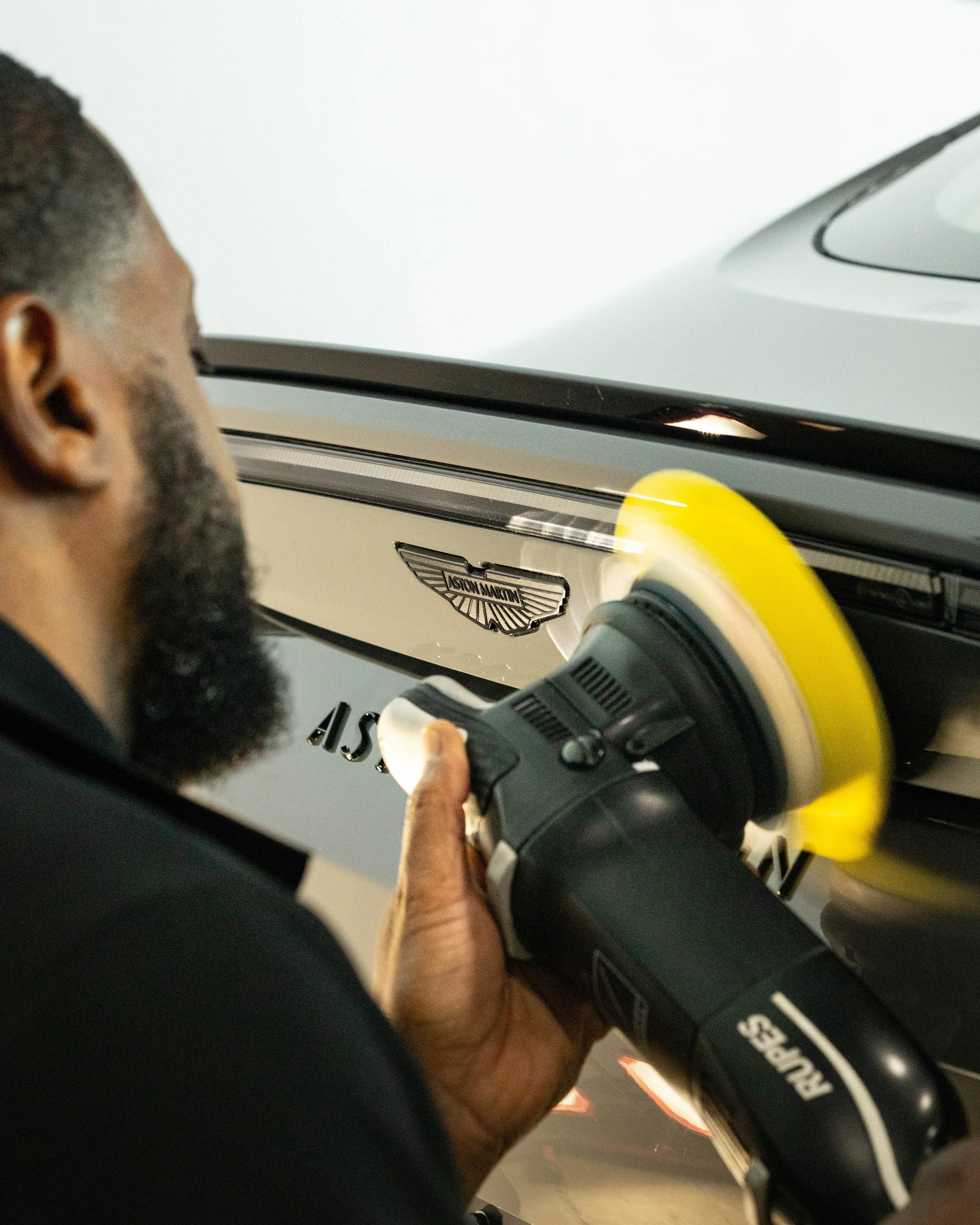 A person polishing a silver Aston Martin car with a yellow polishing machine as the car is recieving a paint correction and ceramic coating