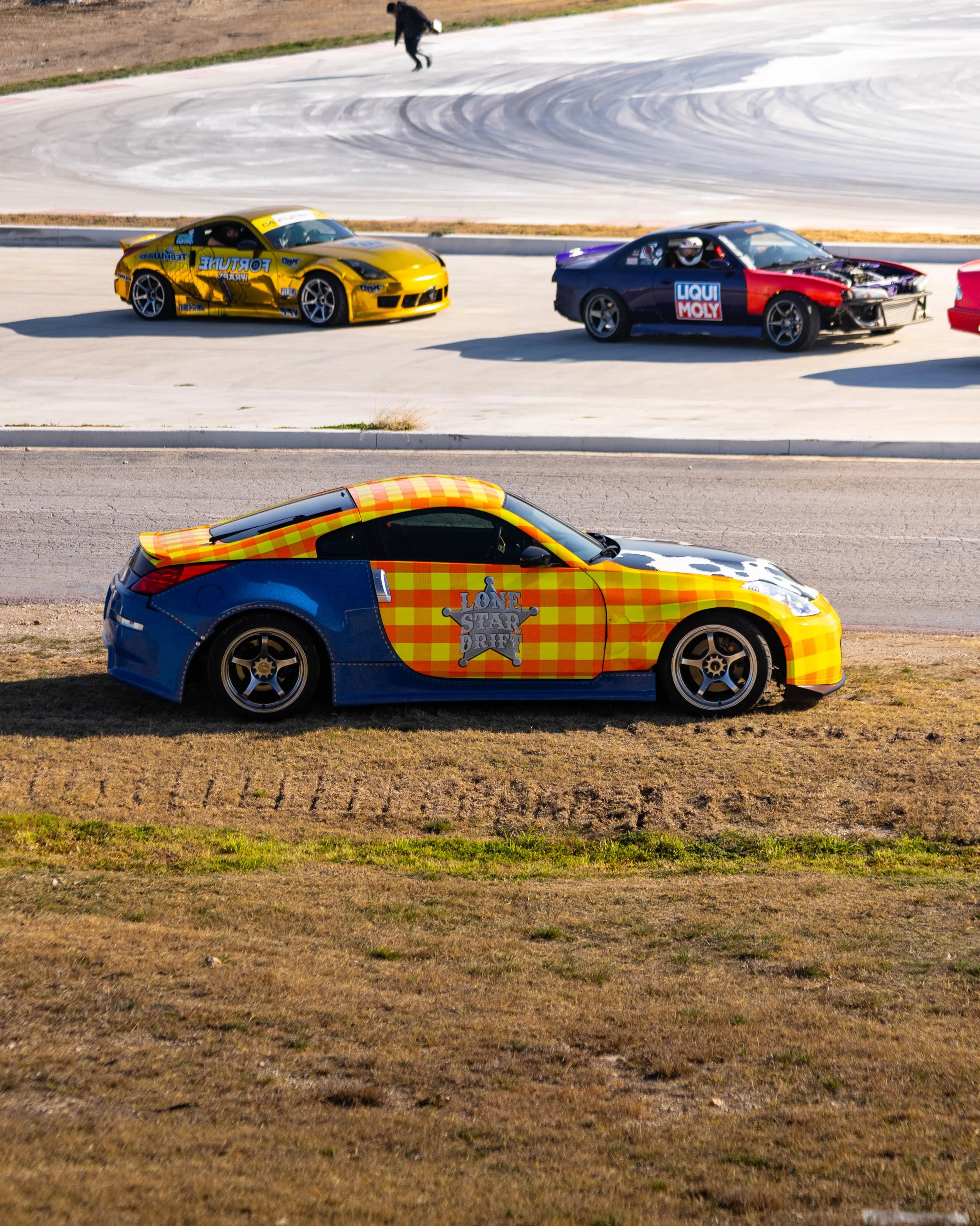 A sports car with a checkered yellow and orange design parked on the grass next to a road, with racing/drifting cars on the track in the background. The car was designed to look like Woody from Toy Story for the owners children to have fun.
