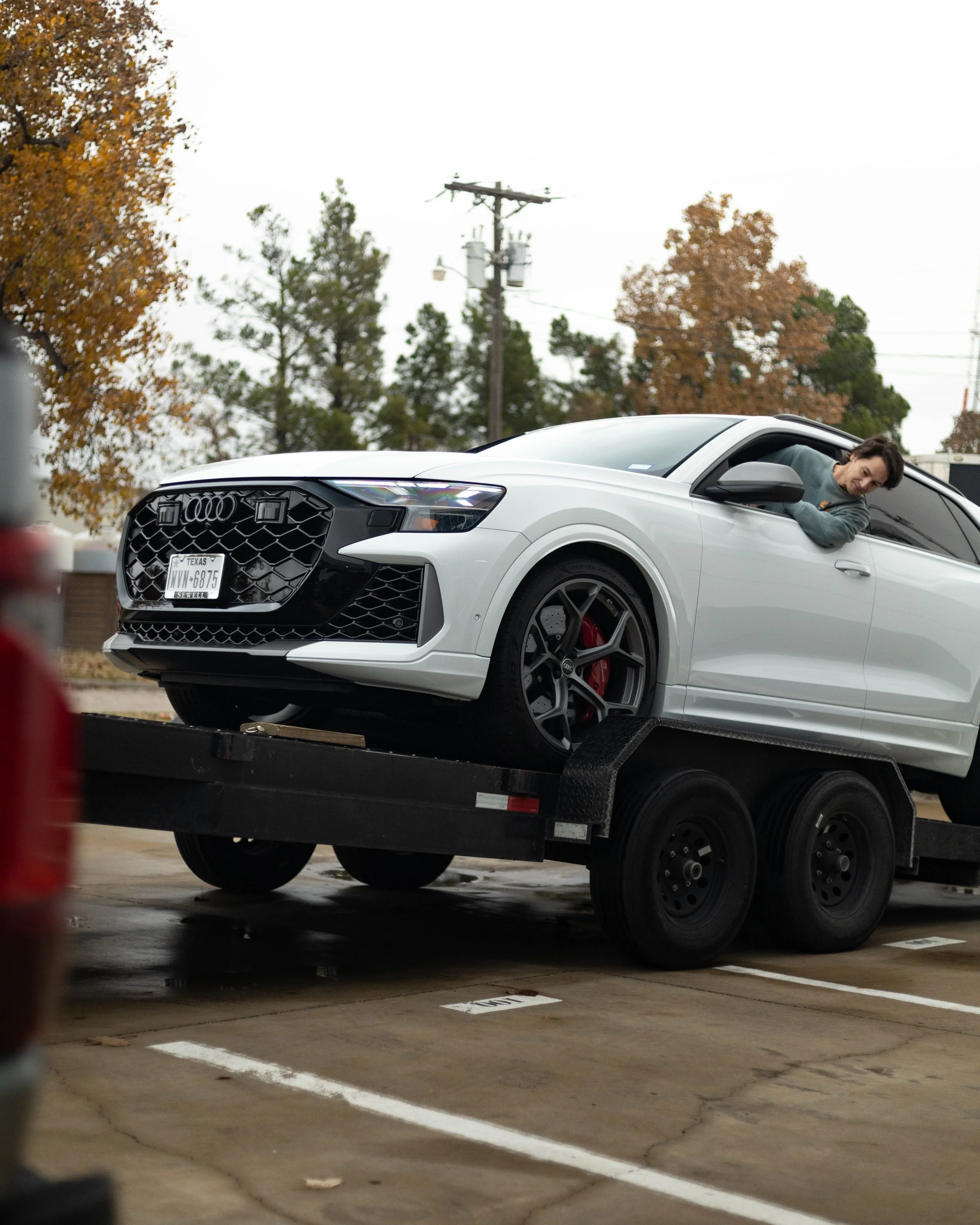 A woman leaning out of the driver's side window of a white Audi SUV parked on a flatbed trailer in a parking lot during autumn.