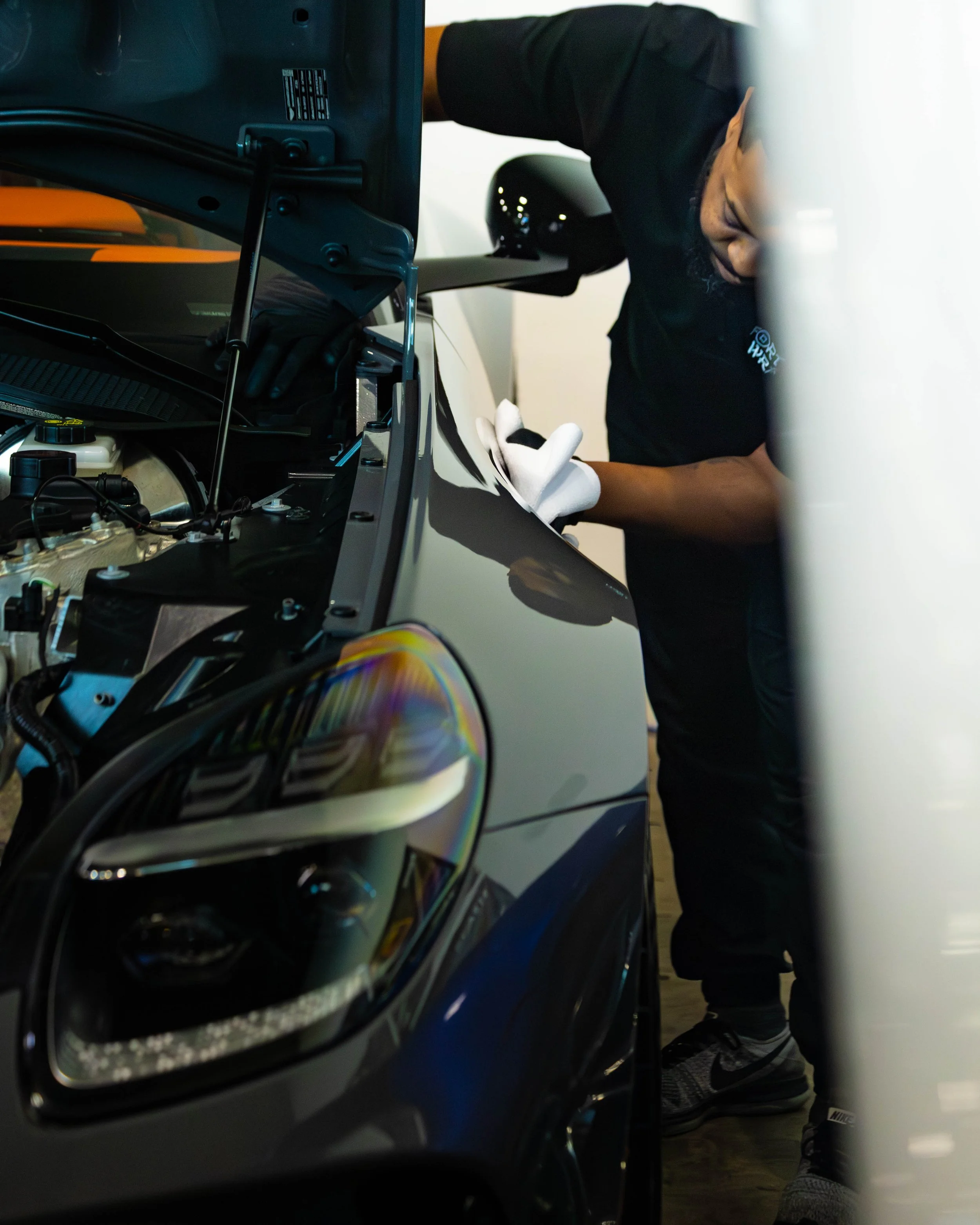 An individual wearing black gloves working on a car's fender with the hood open, using a white cloth to clean or examine the vehicle after installing a protective layer of ceramic coating on the paint.