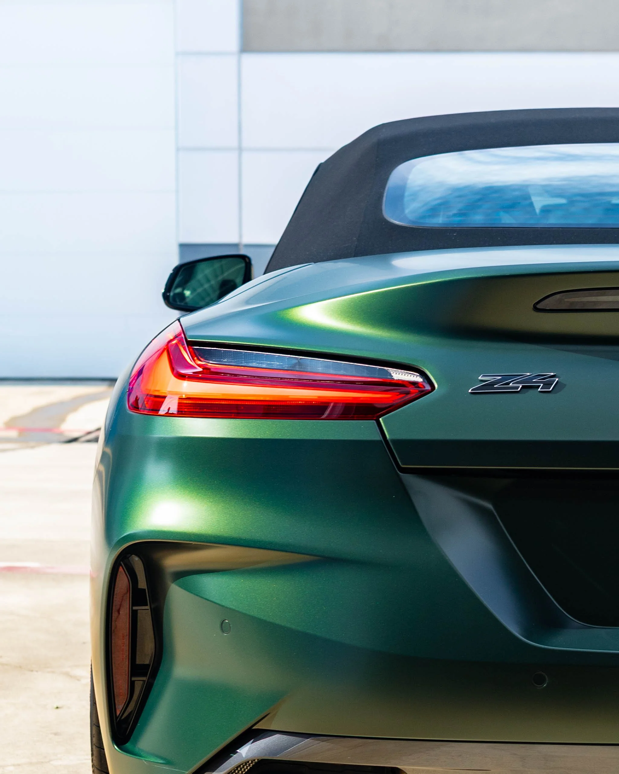 The rear end of a green Mercedes-Benz Z4 convertible with a black soft top, parked outdoors in front of a modern building. this car received matte paint protection film