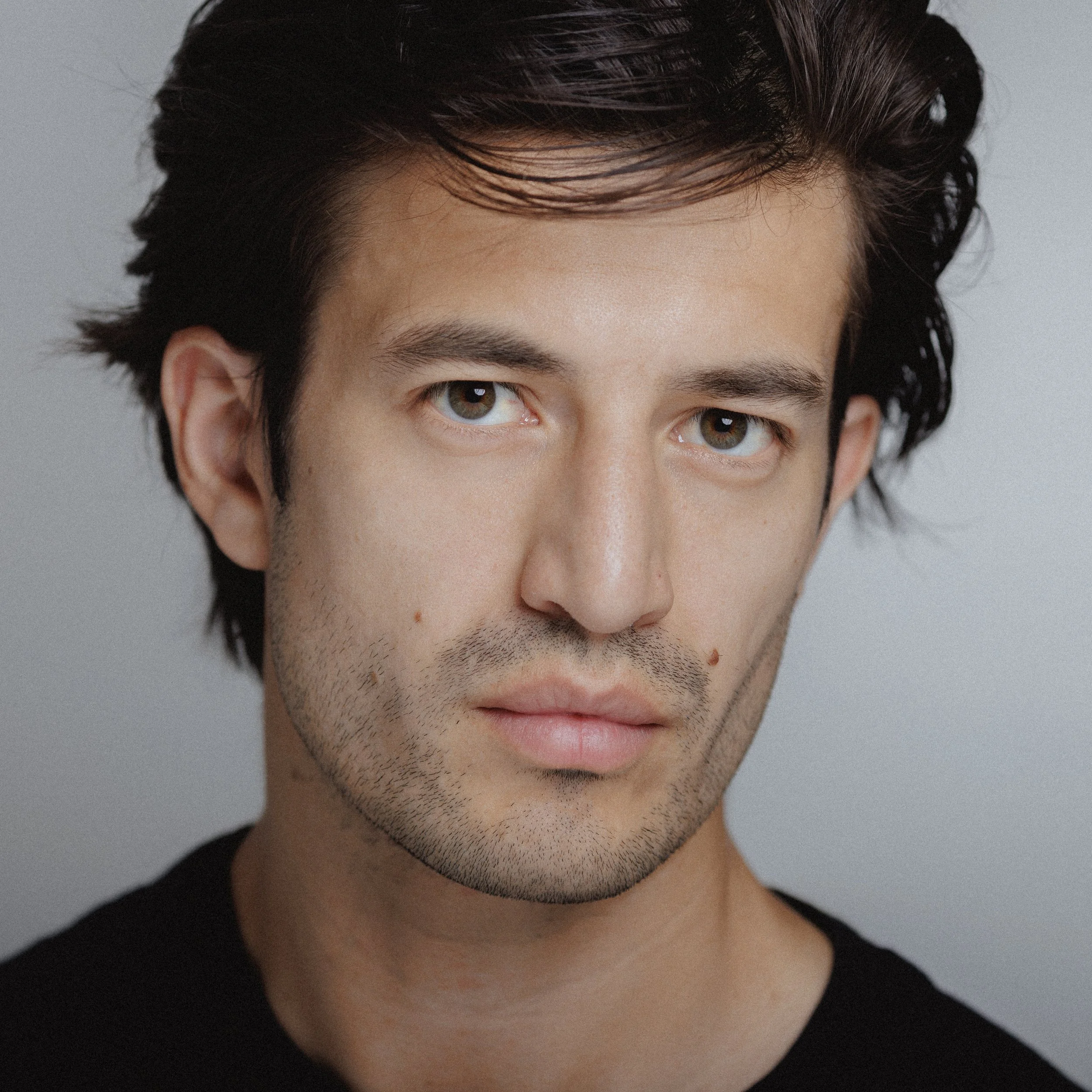 Close-up portrait of a young man with dark brown hair, blue-green eyes, and light facial stubble, looking directly at the camera with a neutral expression.