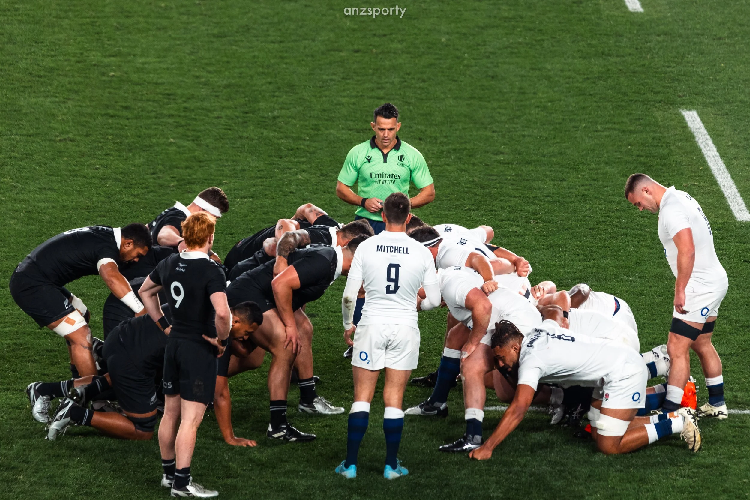 A rugby team engaged in a scrum on the field with players wearing black and white jerseys, and a referee standing behind them.