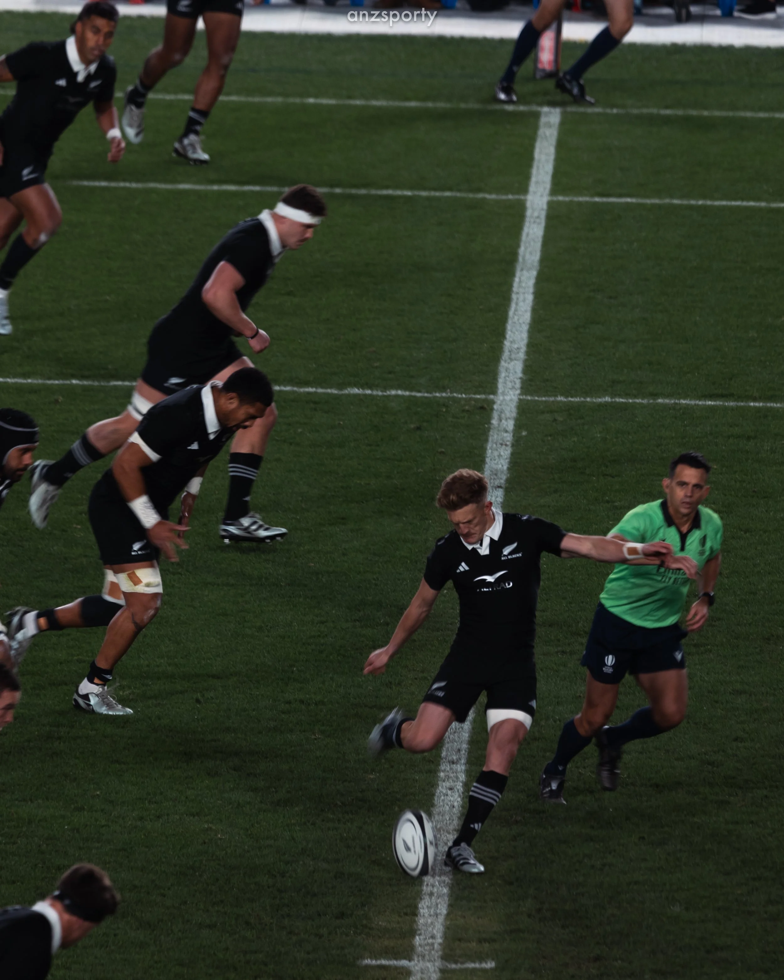 Rugby players in black uniforms on a green field, some running and one kicking the ball, with a referee in a green shirt nearby.