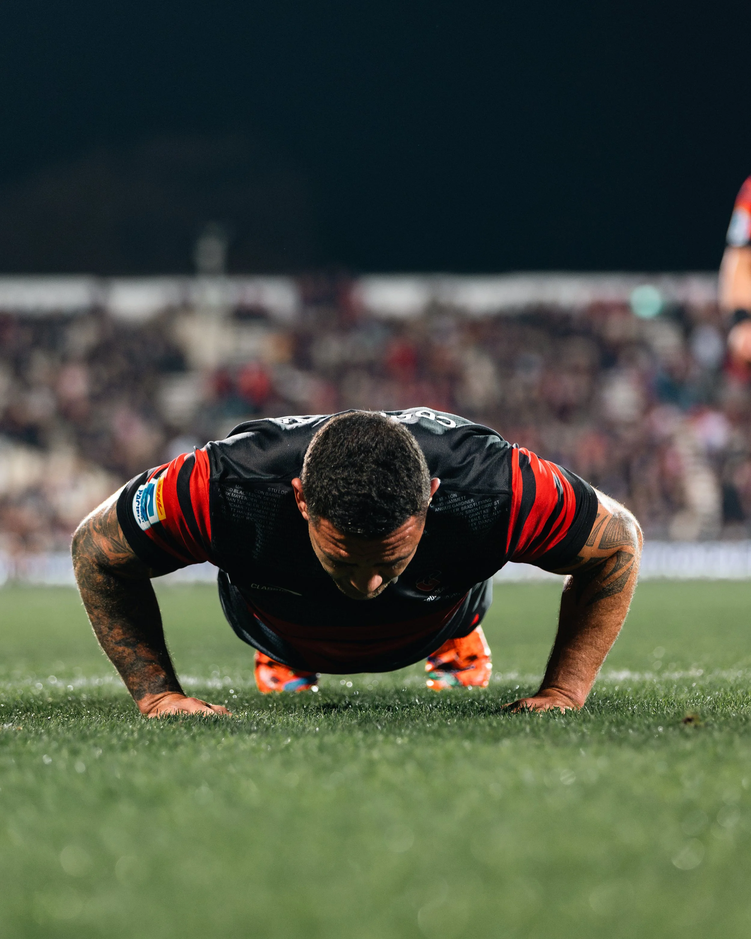 A soccer player in a black and red uniform is doing push-ups on the grass field during a night game with a blurred crowd in the background.