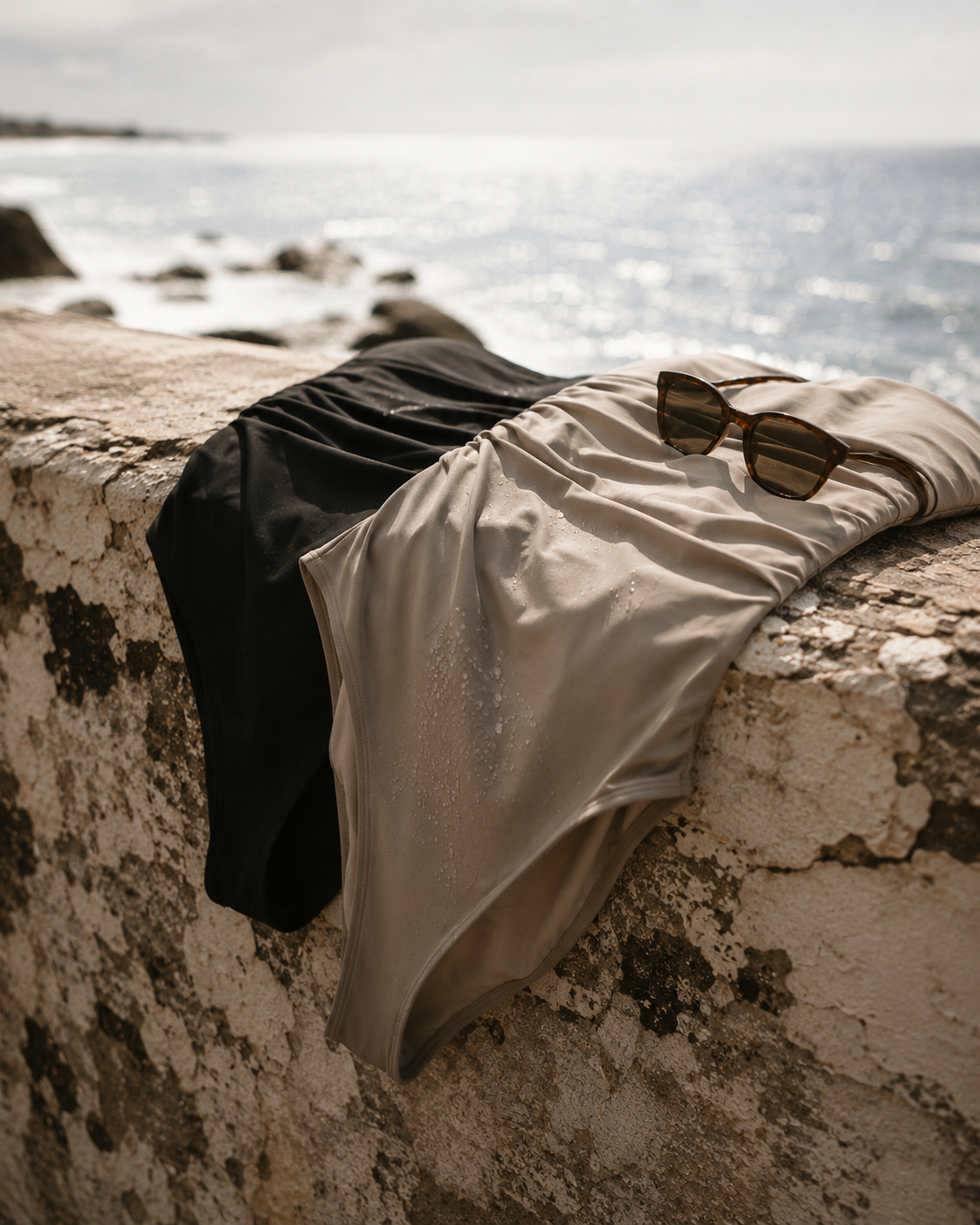 AI campaign visual fashion swimsuit brand: Swimsuit, towels, and sunglasses placed on a stone wall by the ocean with rocks and water in the background.