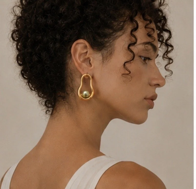 AI campaign visual jewellery  
A woman with curly hair wearing gold earrings with a pearl inside, facing sideways.