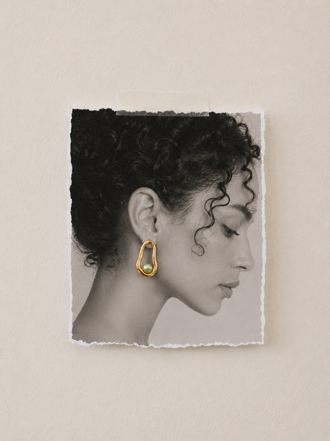 AI campaign visual jewellery Black and white portrait of a woman with curly hair, wearing a gold earring with a green pearl, taped to a beige wall.