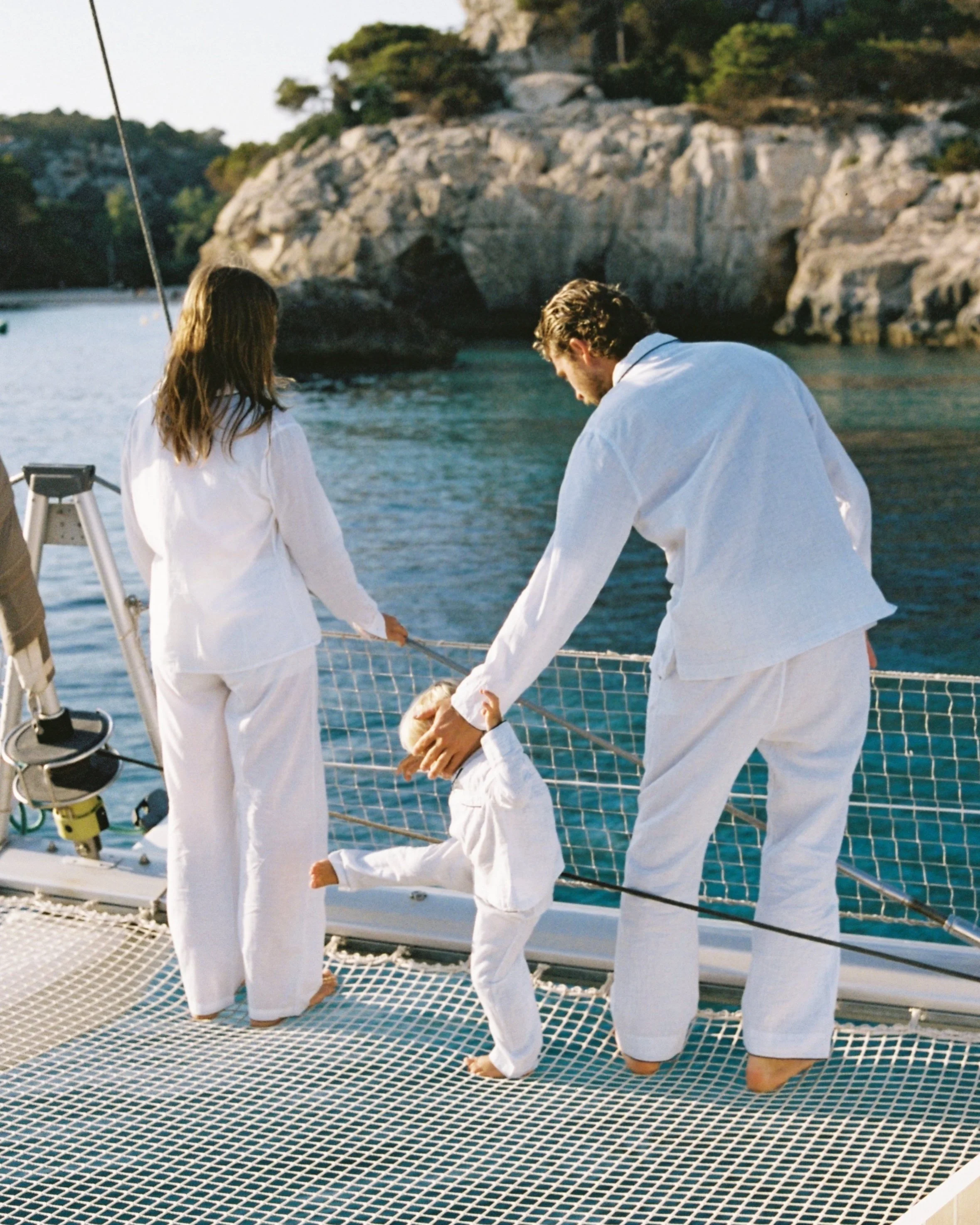 A family on a sailboat near rocky coastline, with a man and woman holding hands and a small child in white outfits.