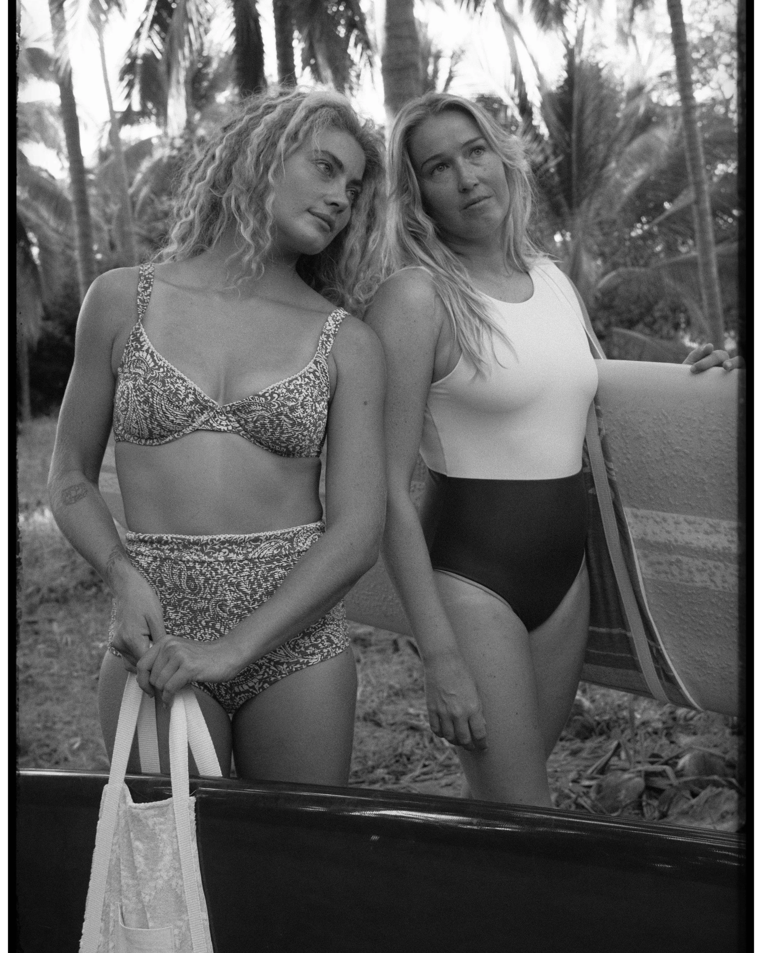 Two women in swimsuits standing outdoors under palm trees, with one holding a tote bag and a surfboard.