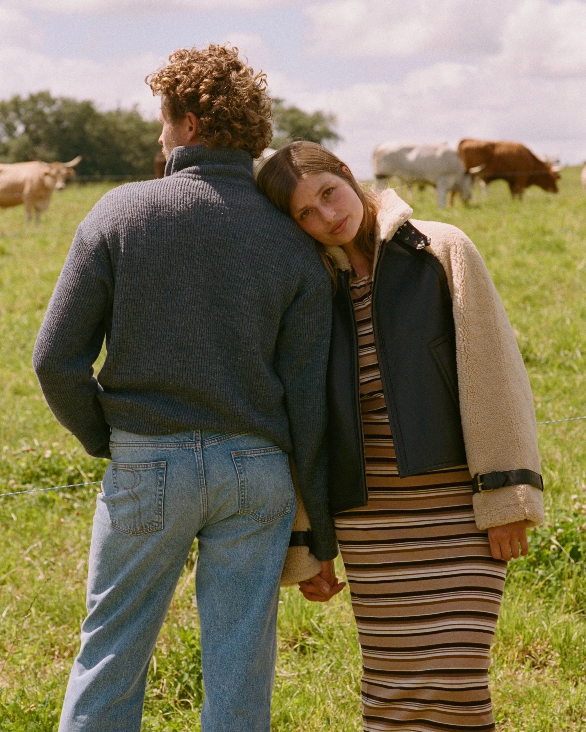 A woman with brown hair leaning her head on a man's shoulder in a field with cows in the background.