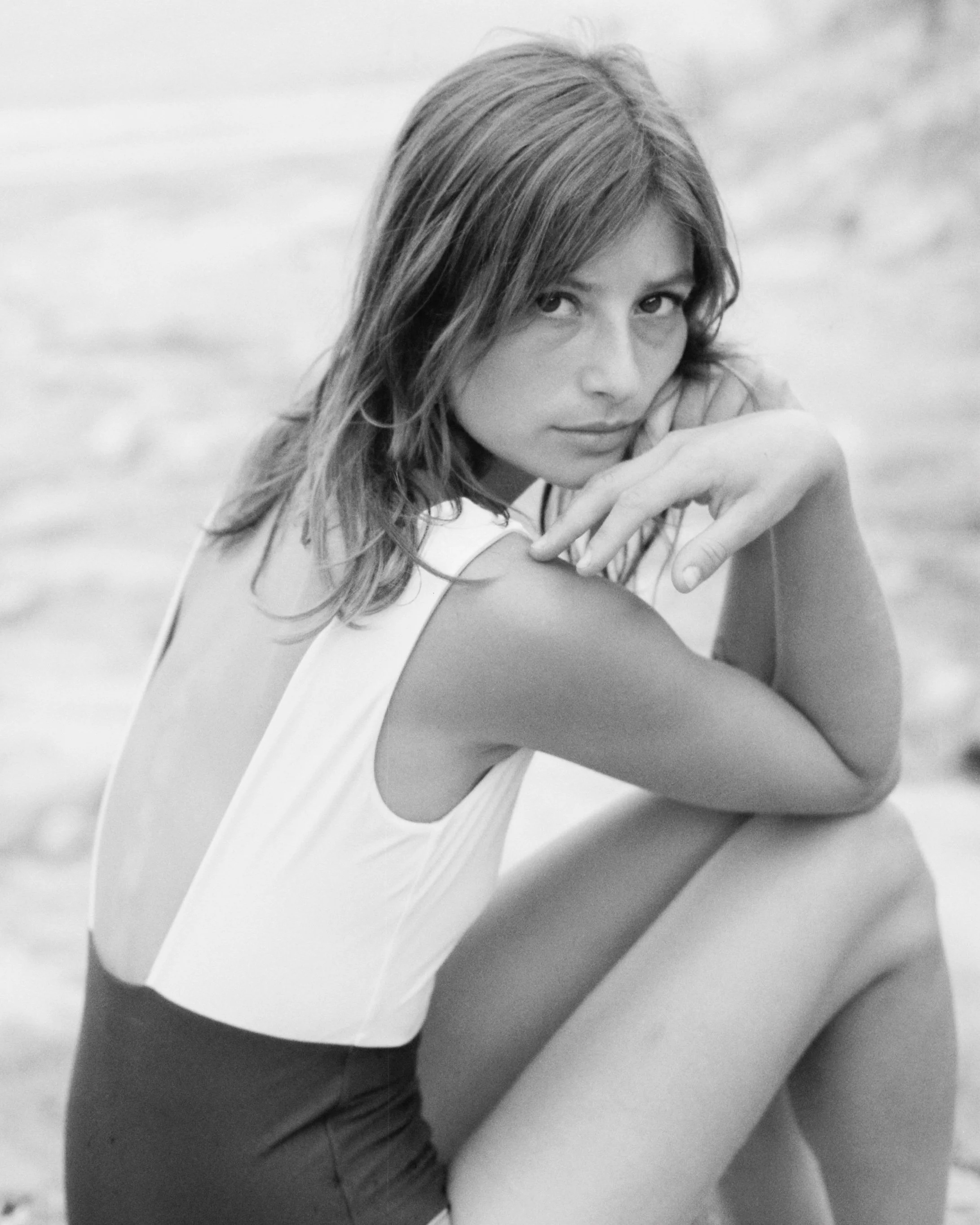 A black and white photo of a woman with shoulder-length hair sitting on a beach, looking at the camera with her chin resting on her hand.