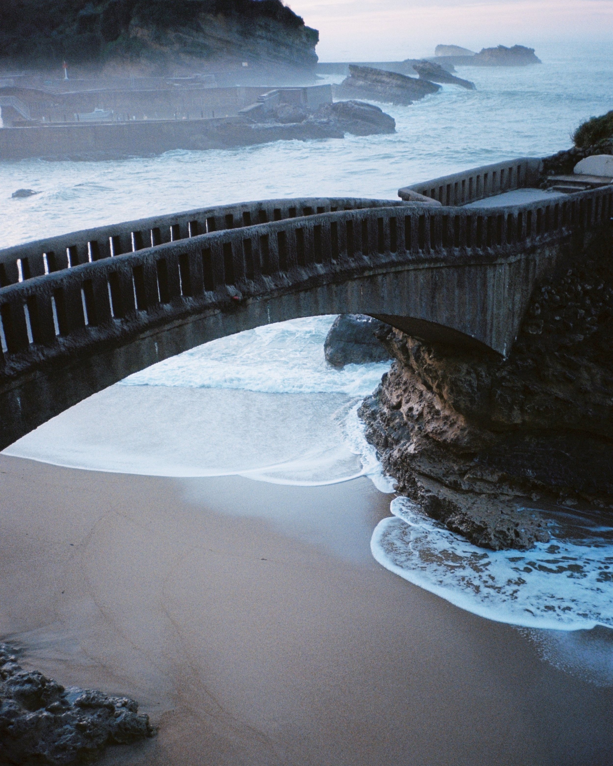 A bridge over a rocky cliff, leading to a sandy beach with ocean waves crashing against rocks, during dusk or dawn.
