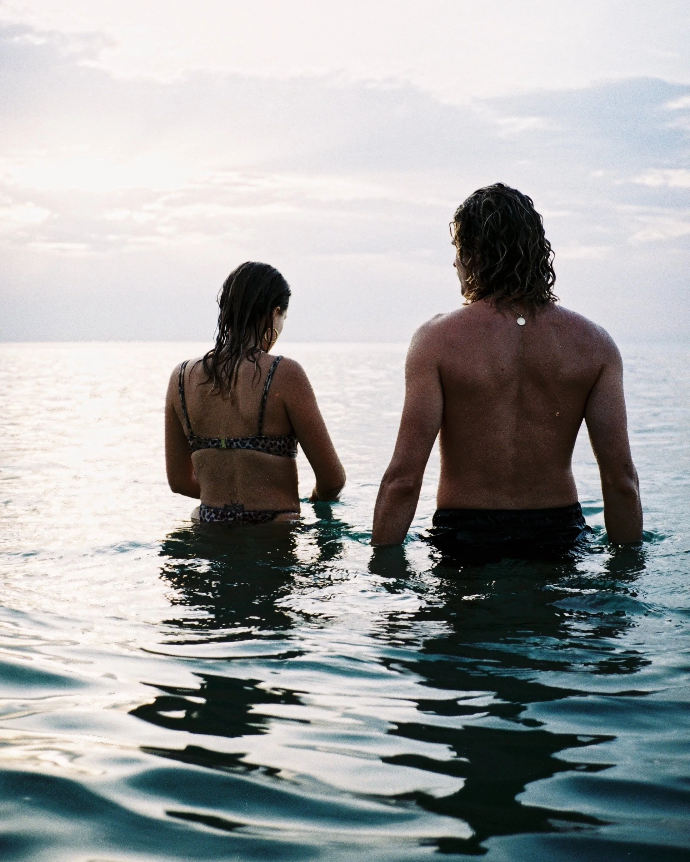 Two people with long hair standing in the ocean during sunset, viewed from behind.