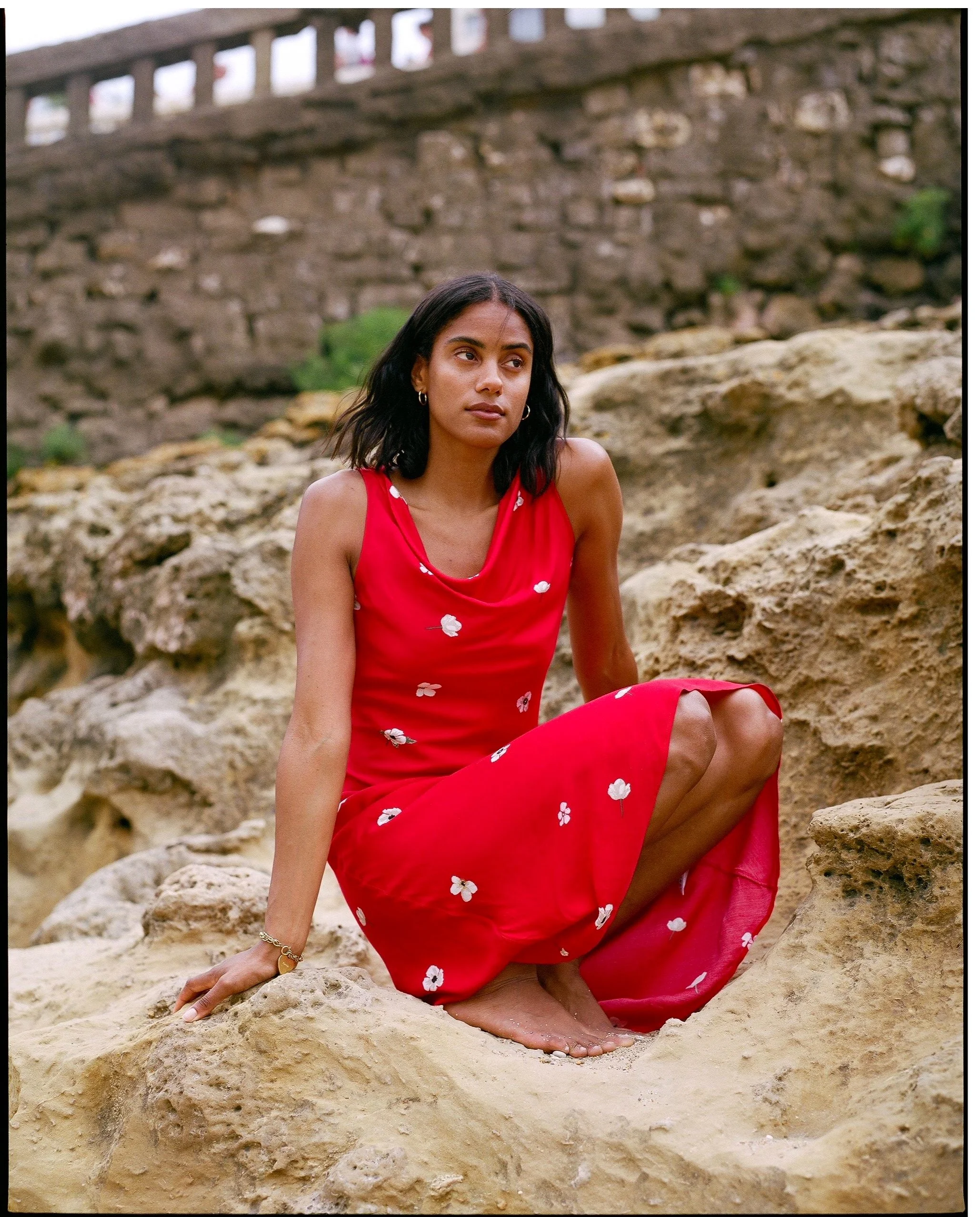 A woman in a red dress with white floral patterns sitting barefoot on rocks near a stone wall.