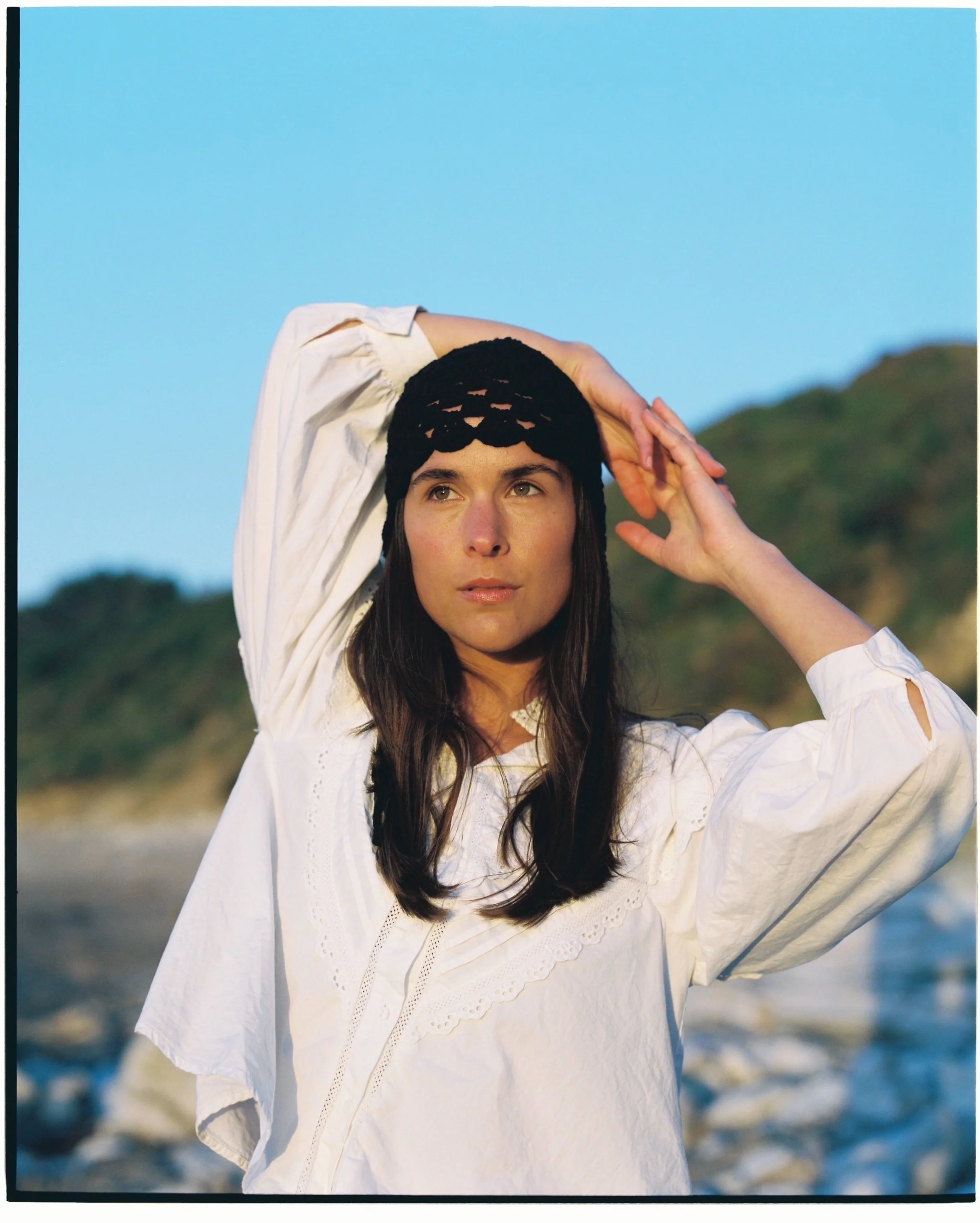 A woman stands outdoors at the beach with a background of green hills and blue sky, wearing a white blouse and a black headscarf, posing with one hand on her head.