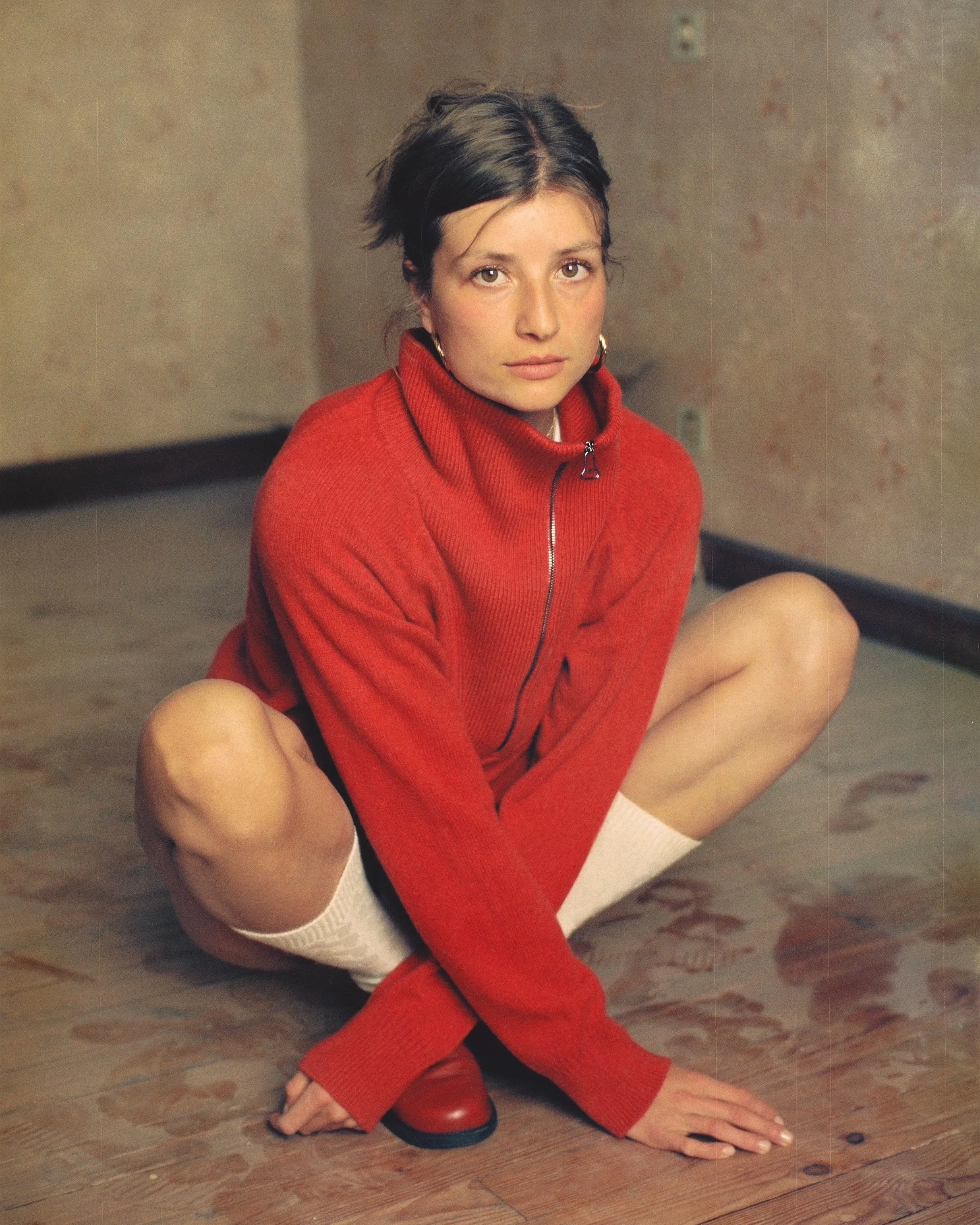 A woman with short dark hair sitting on a wooden floor, wearing a red zip-up sweater, white knee-high socks, and red shoes, with a neutral expression.