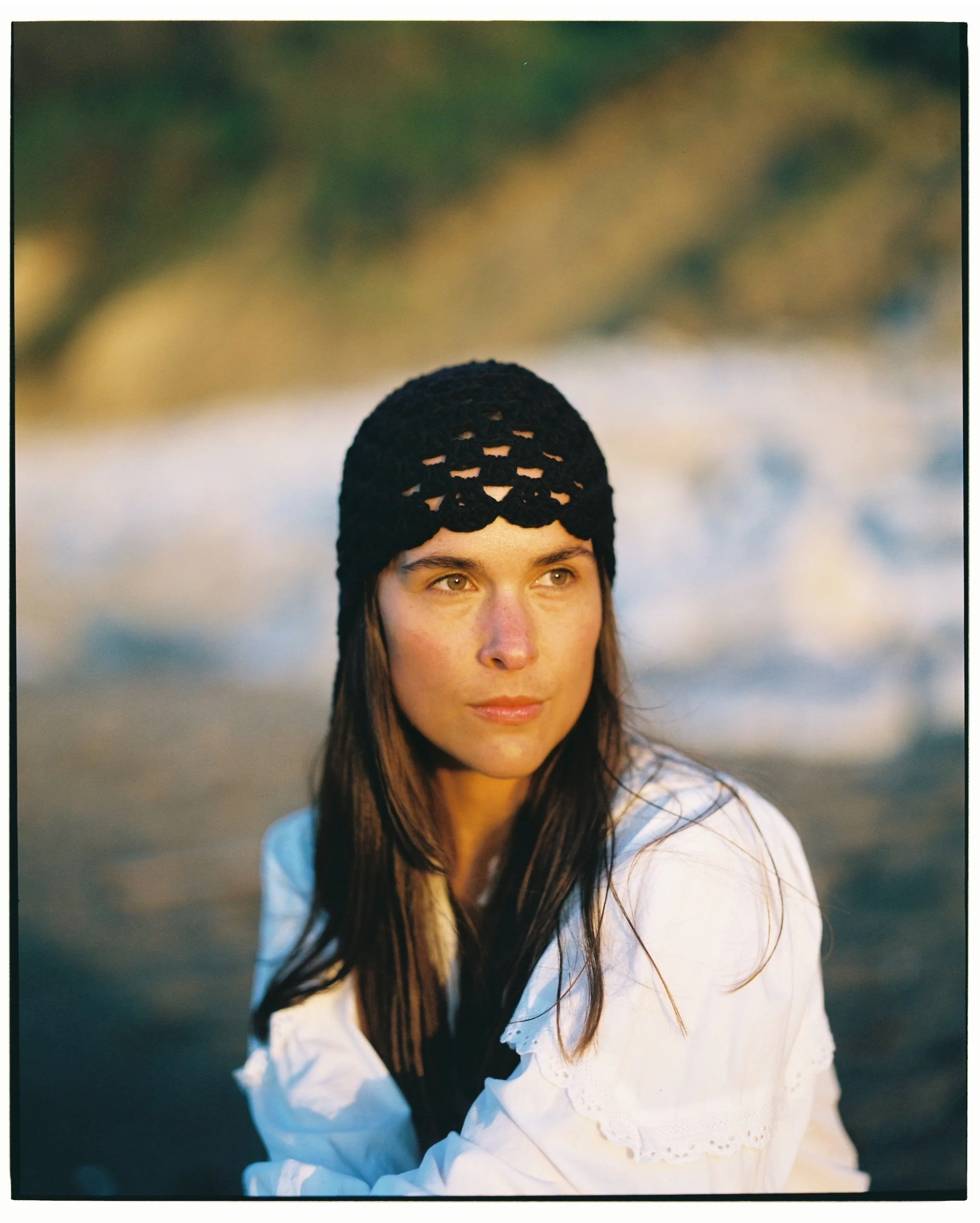 A woman with long brown hair wearing a black crocheted hat and a white blouse, outdoors near water with blurred natural background.