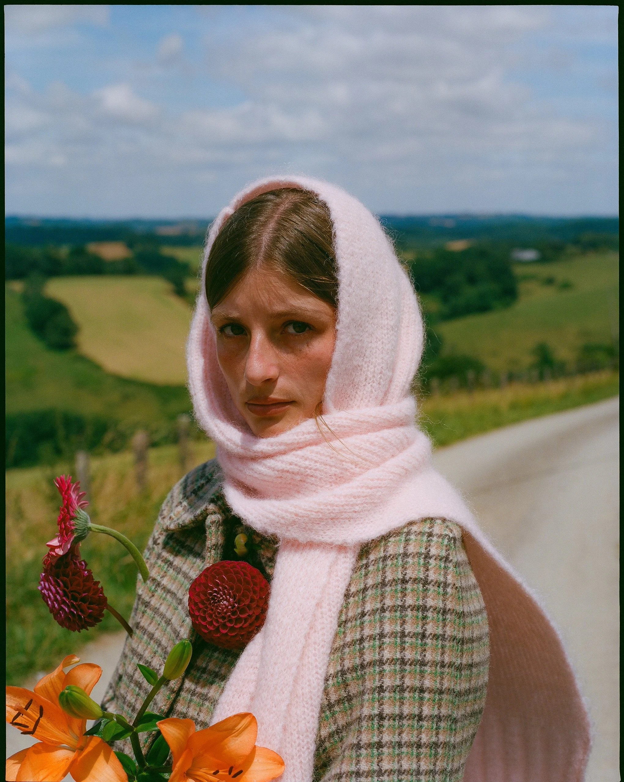 A woman wearing a pink headscarf and plaid coat holding a bouquet of orange and red flowers, standing outdoors with a scenic countryside background of green fields and a cloudy sky.