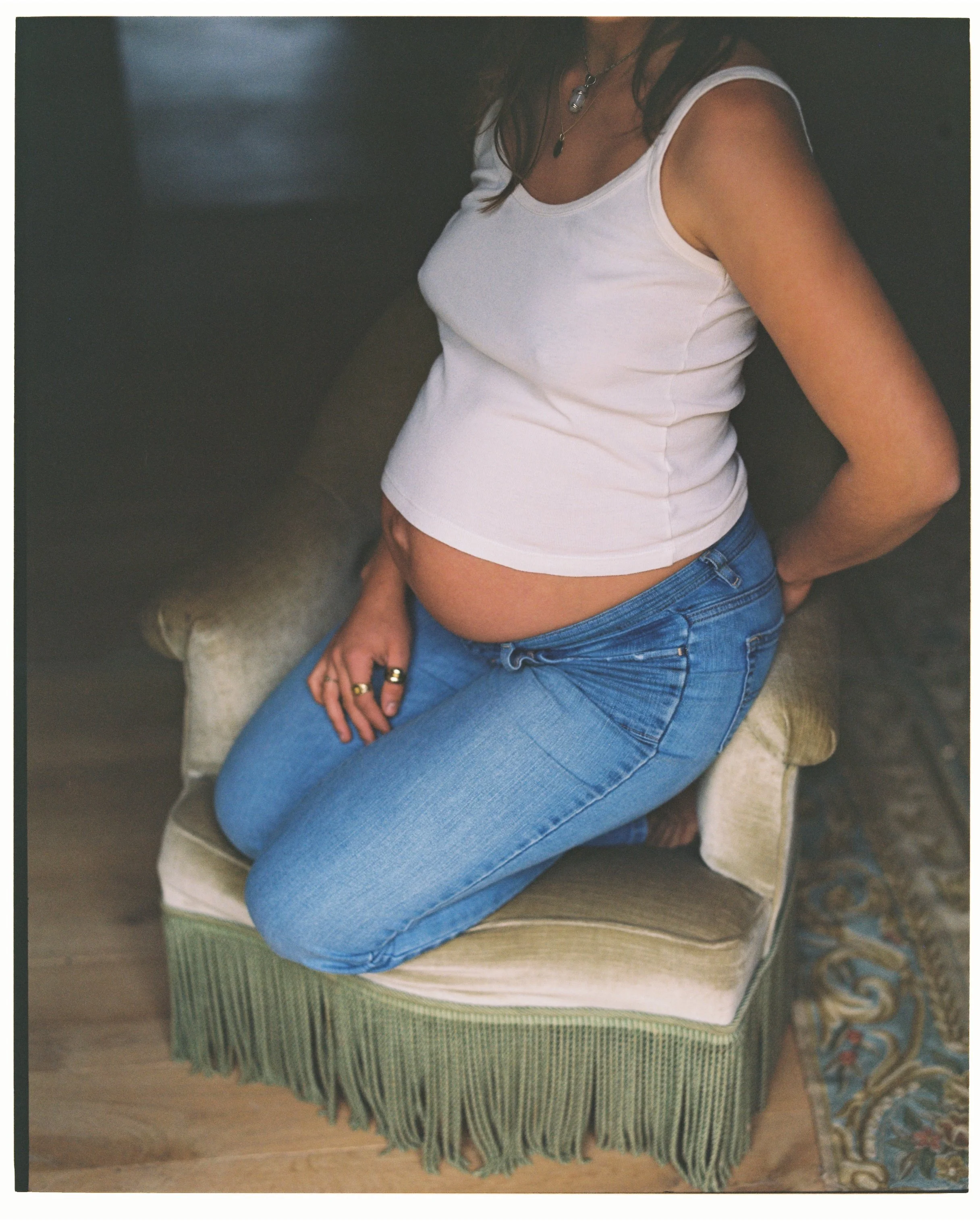 A woman in a white tank top and blue jeans kneeling on a vintage green armchair.