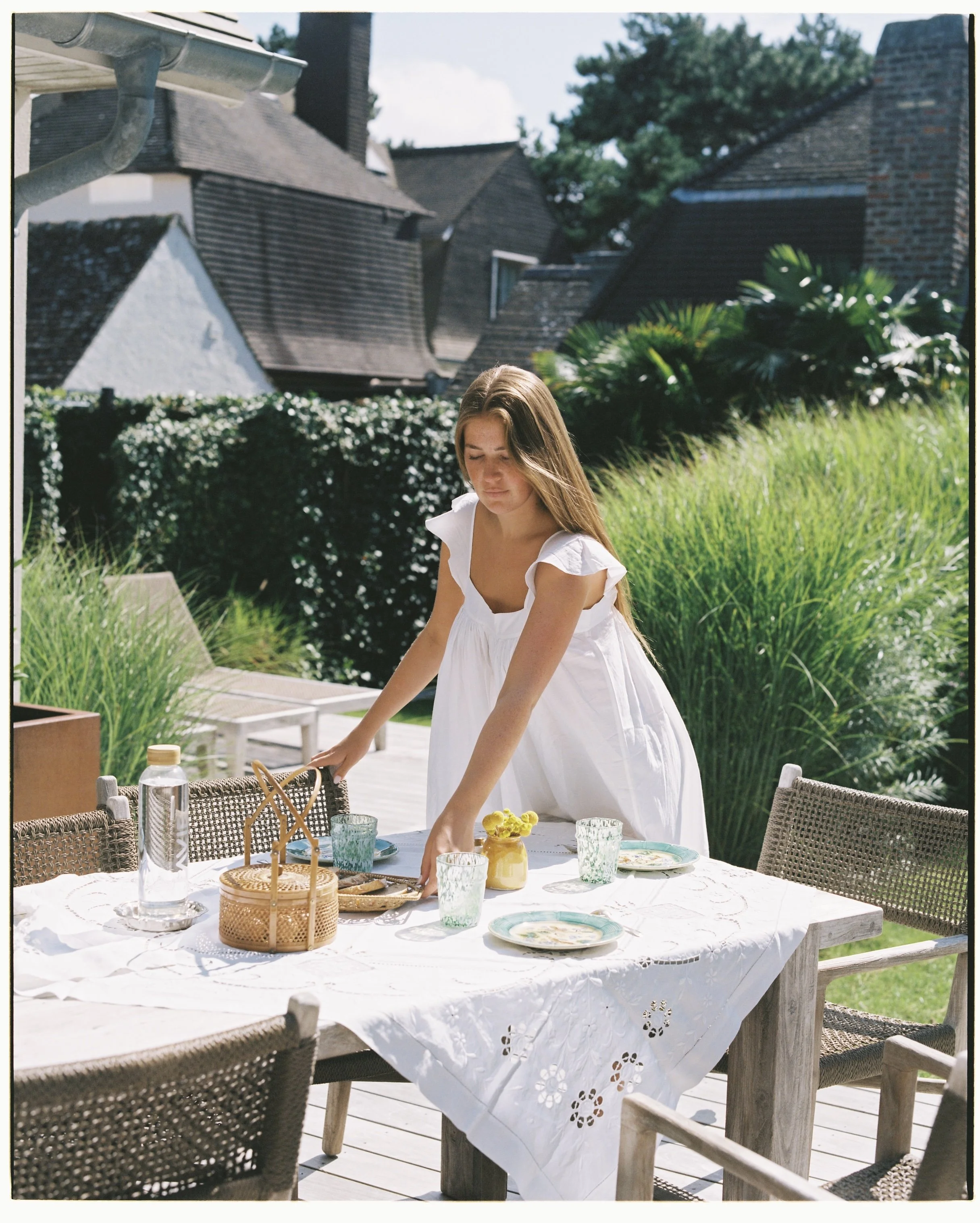 A woman in a white dress setting an outdoor dining table with plates, glasses, a water bottle, and a small yellow potted plant on a sunny day.