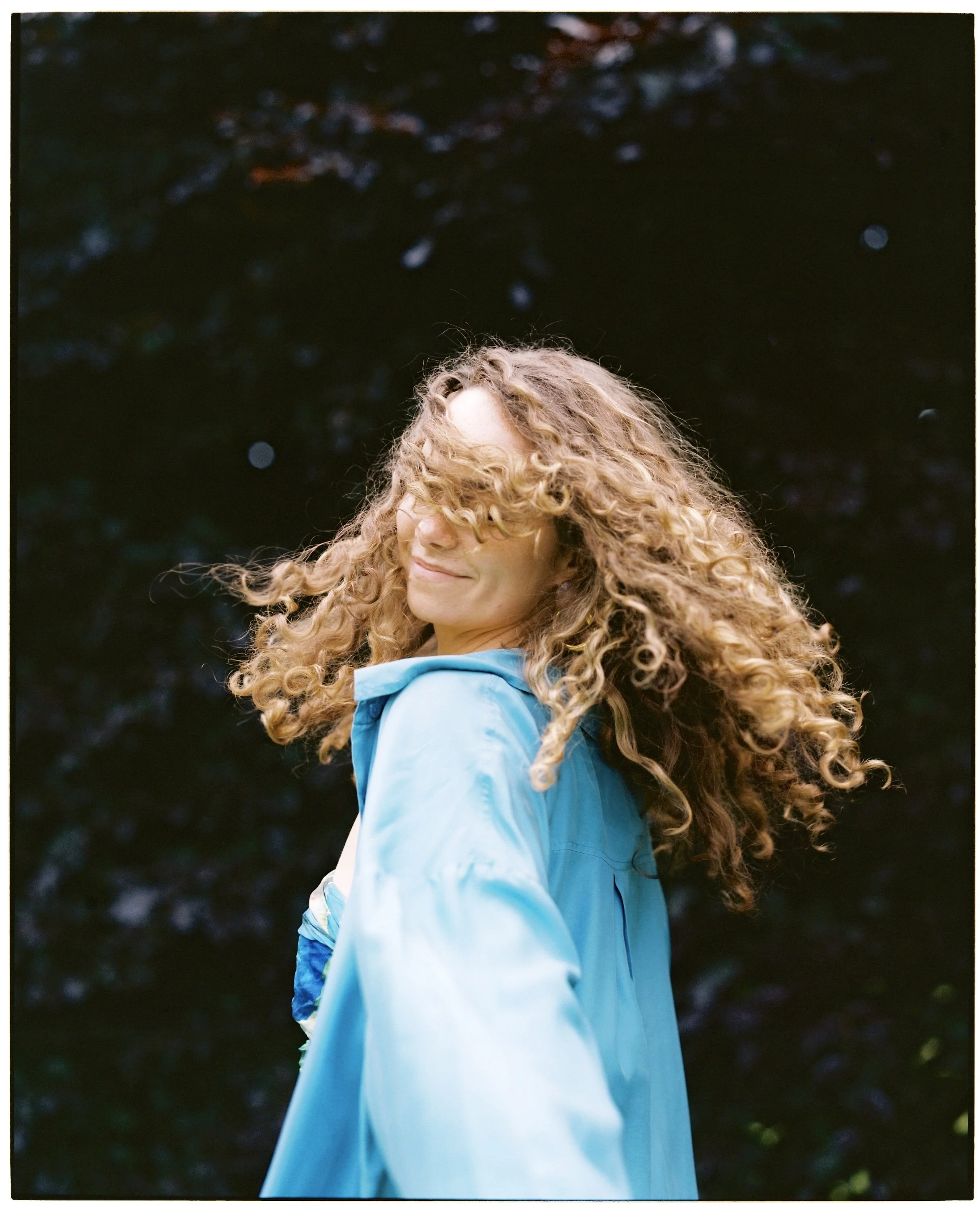 Woman with curly hair smiling and turning her head to the side, wearing a light blue jacket, outdoors with dark foliage background.