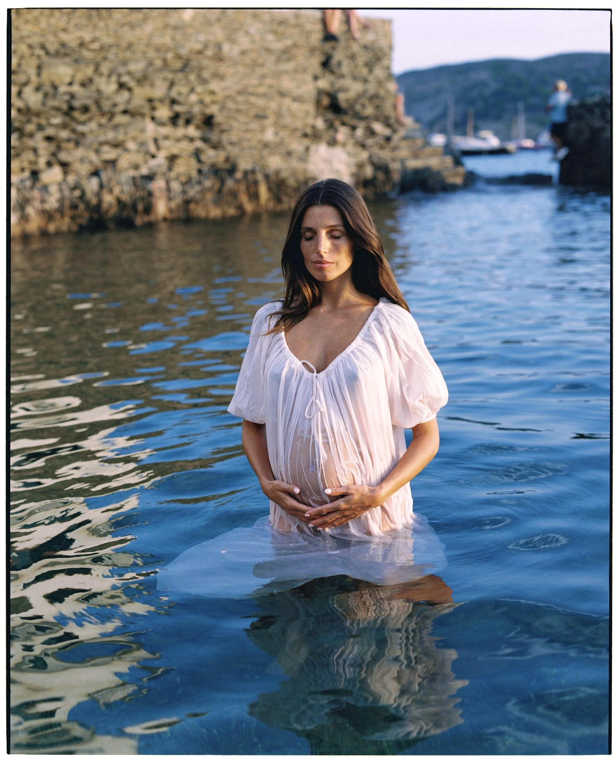 A pregnant woman in a white dress stands in a body of water with her hands on her belly, eyes closed, and a serene expression.