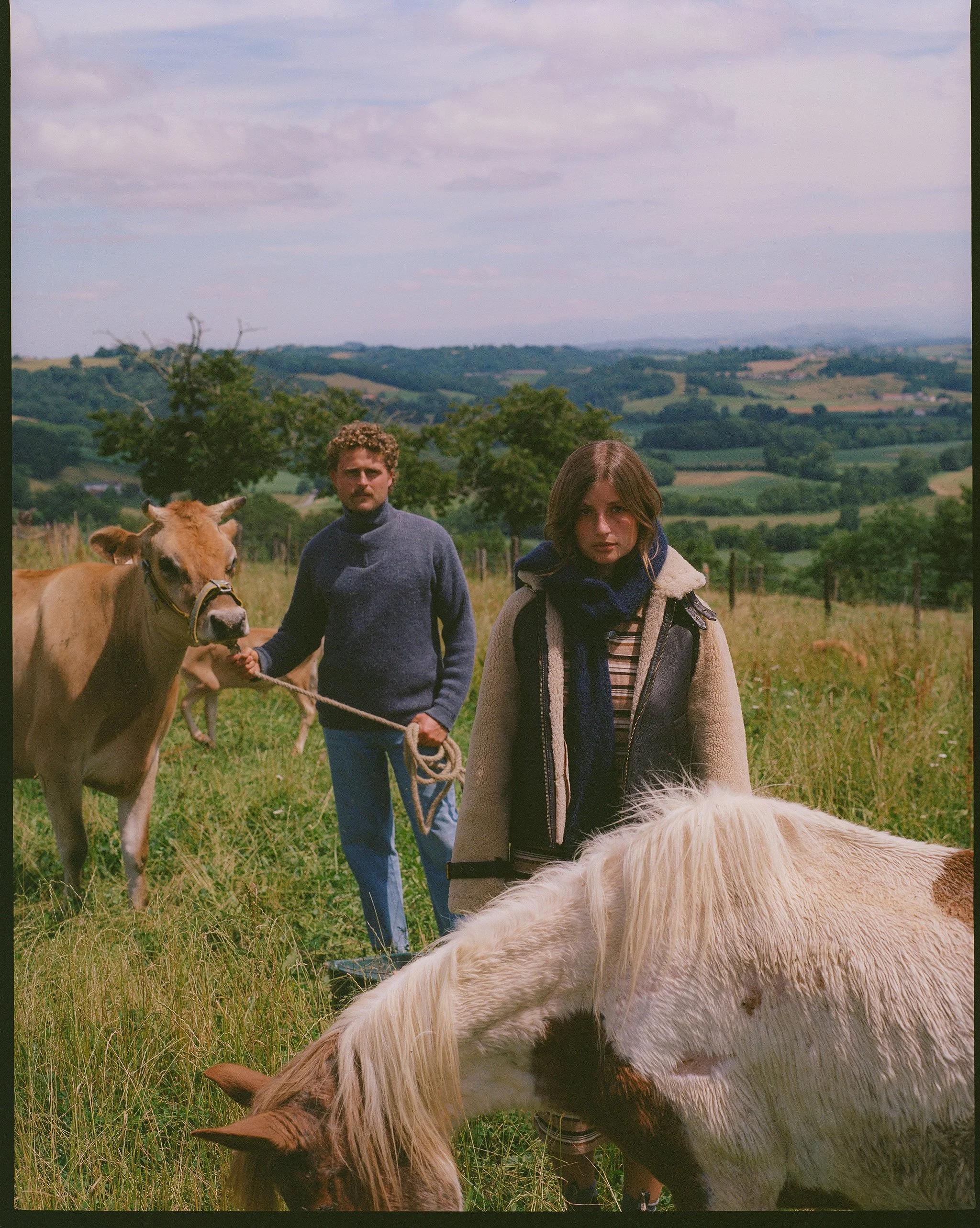 A young woman and a young man in a field with horses and cows, with rolling hills and a cloudy sky in the background.