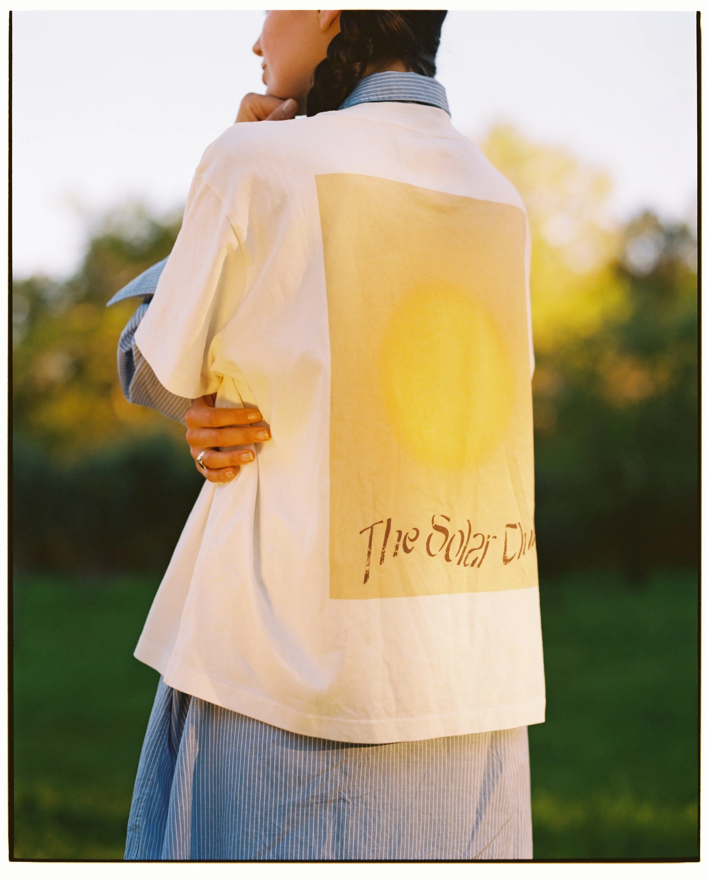 Back of a person wearing a white t-shirt with a yellow square and text that reads "The Solar Dip" in a green outdoor setting with trees in the background.