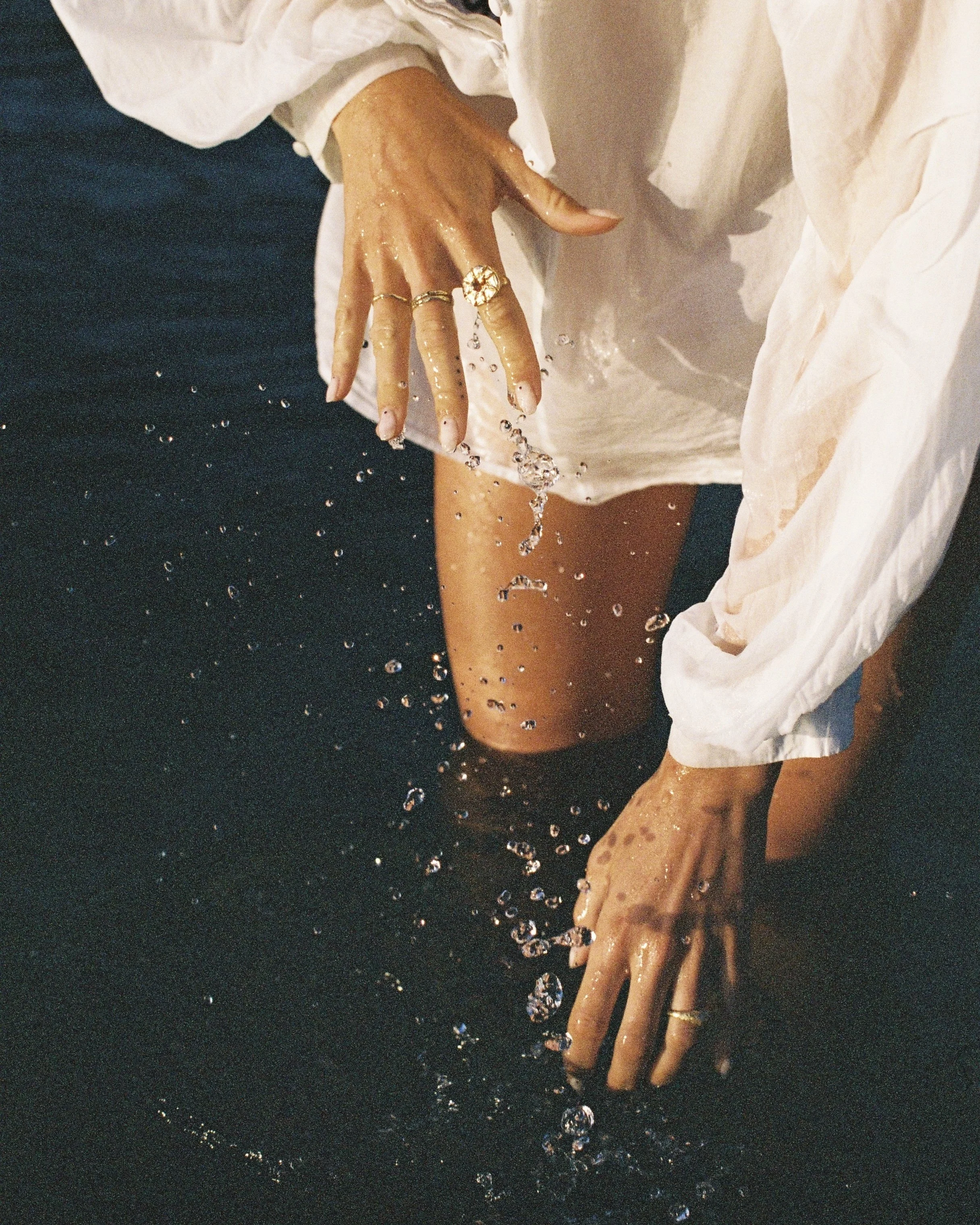 Person wearing rings is kneeling in water, splashing with hands, wearing a white shirt with rolled-up sleeves.