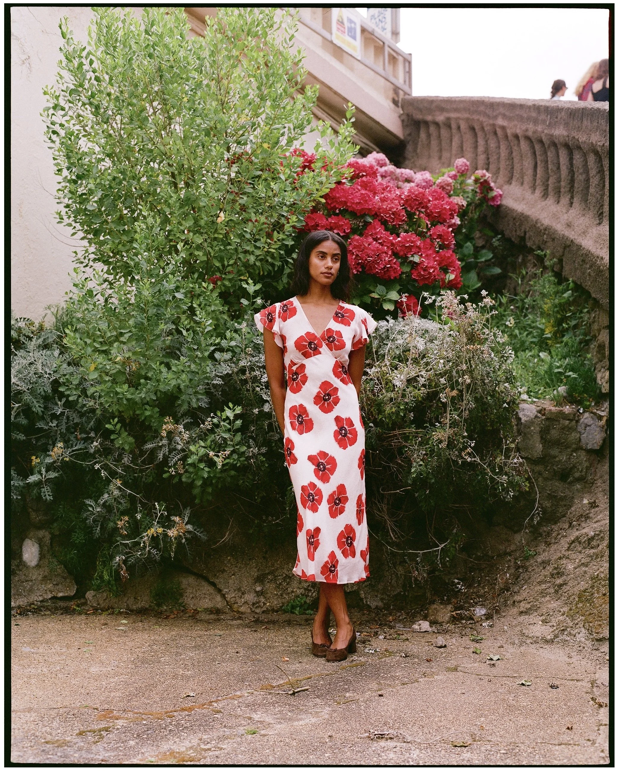 A woman in a white dress with red floral patterns standing in front of green foliage and pink flowers on a stone wall, with a stone staircase and people in the background.