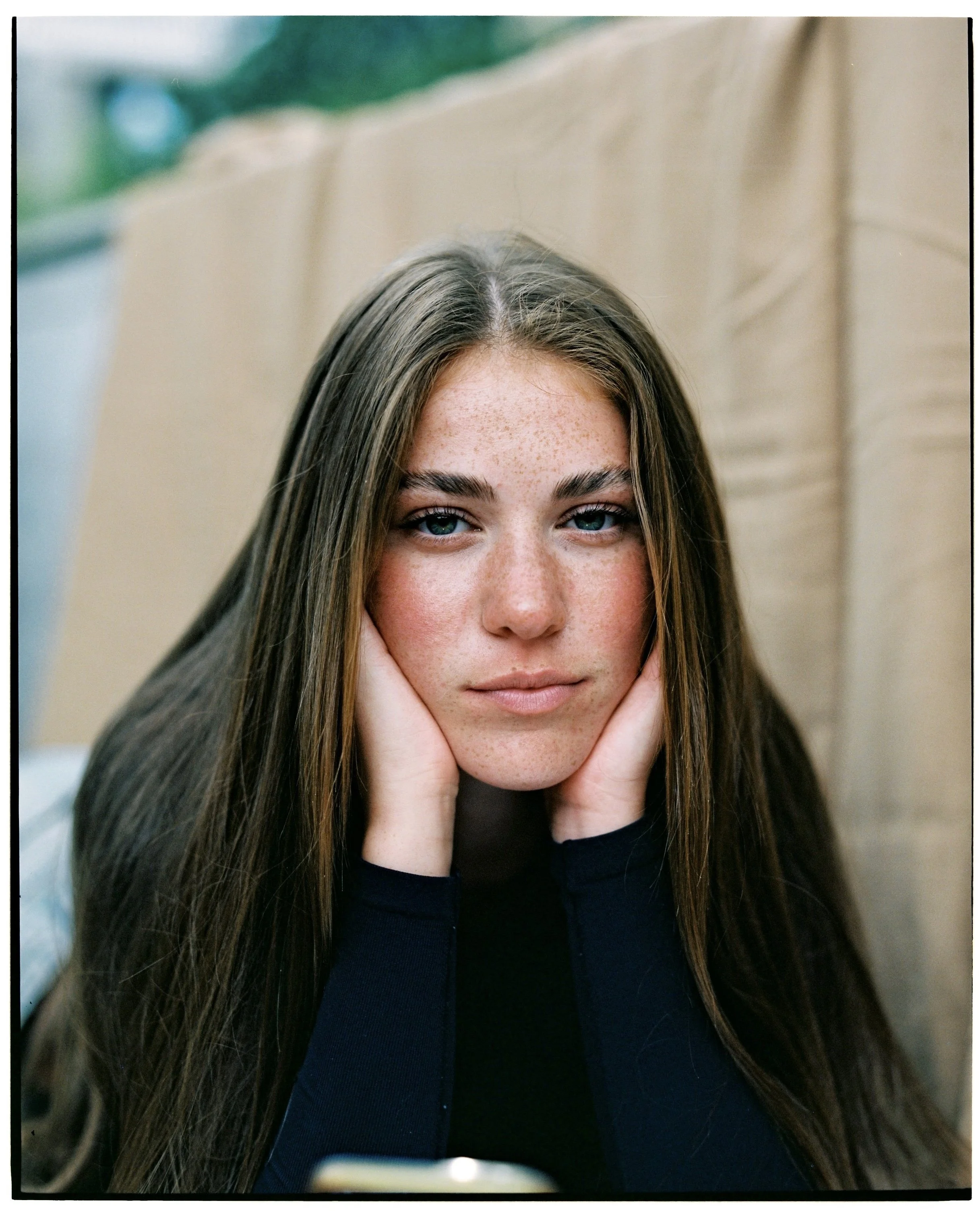 Close-up of a young woman with long brown hair, blue eyes, and freckles, resting her face in her hands while sitting indoors.