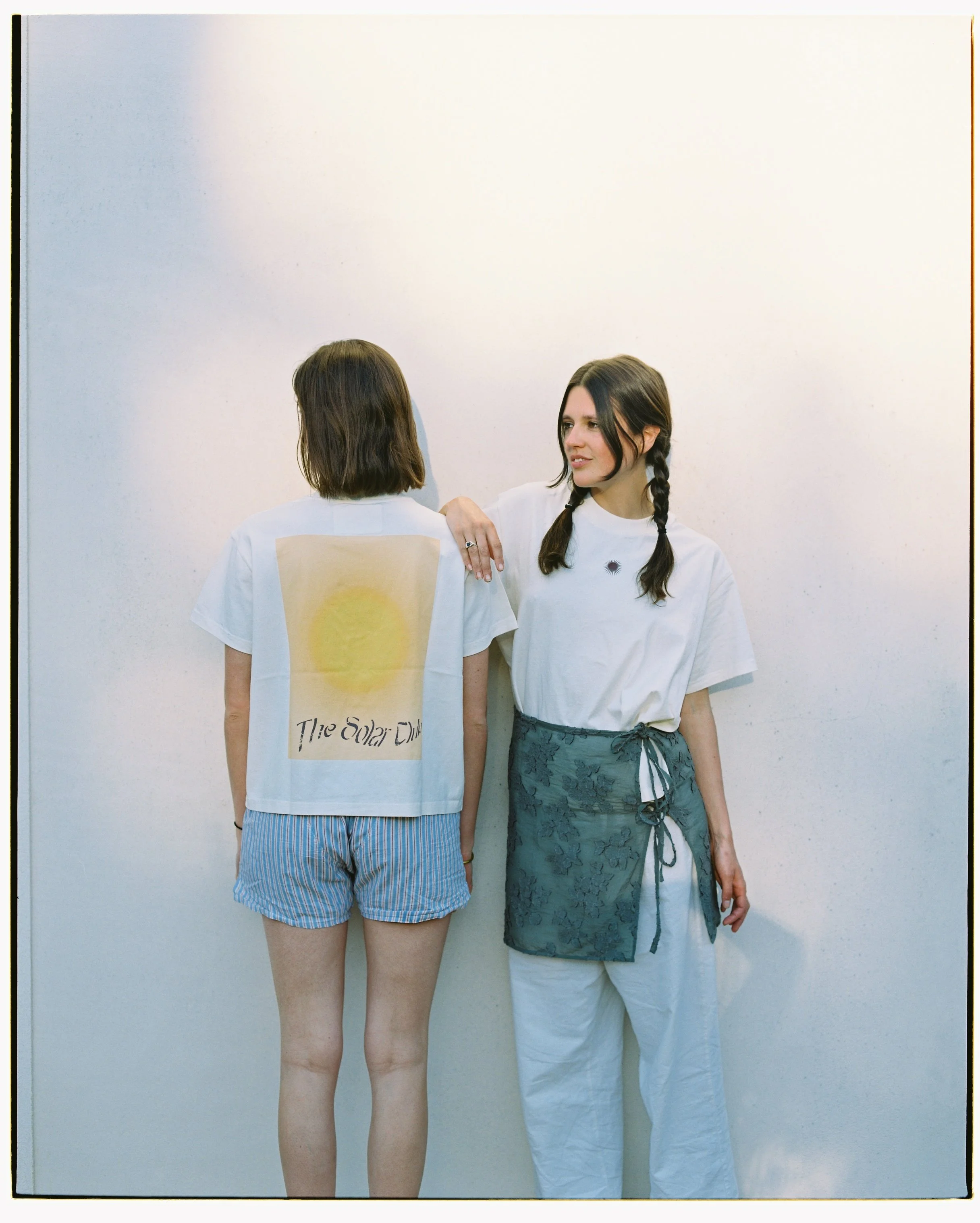 Two young women with dark hair and casual clothing stand against a plain white wall, one facing away and one facing sideways, engaged in a conversation.