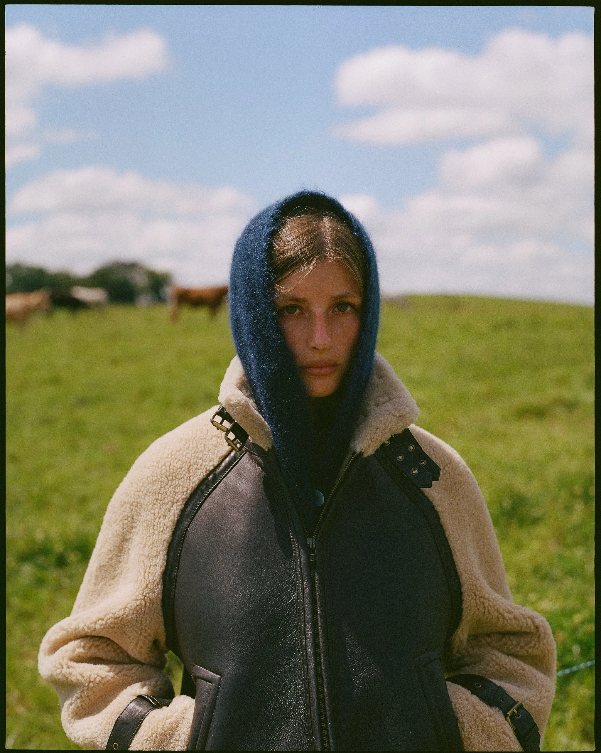 A young woman with a serious expression, wearing a navy hood and a beige and black jacket, stands in a grassy field with cows and a blue sky with clouds in the background.