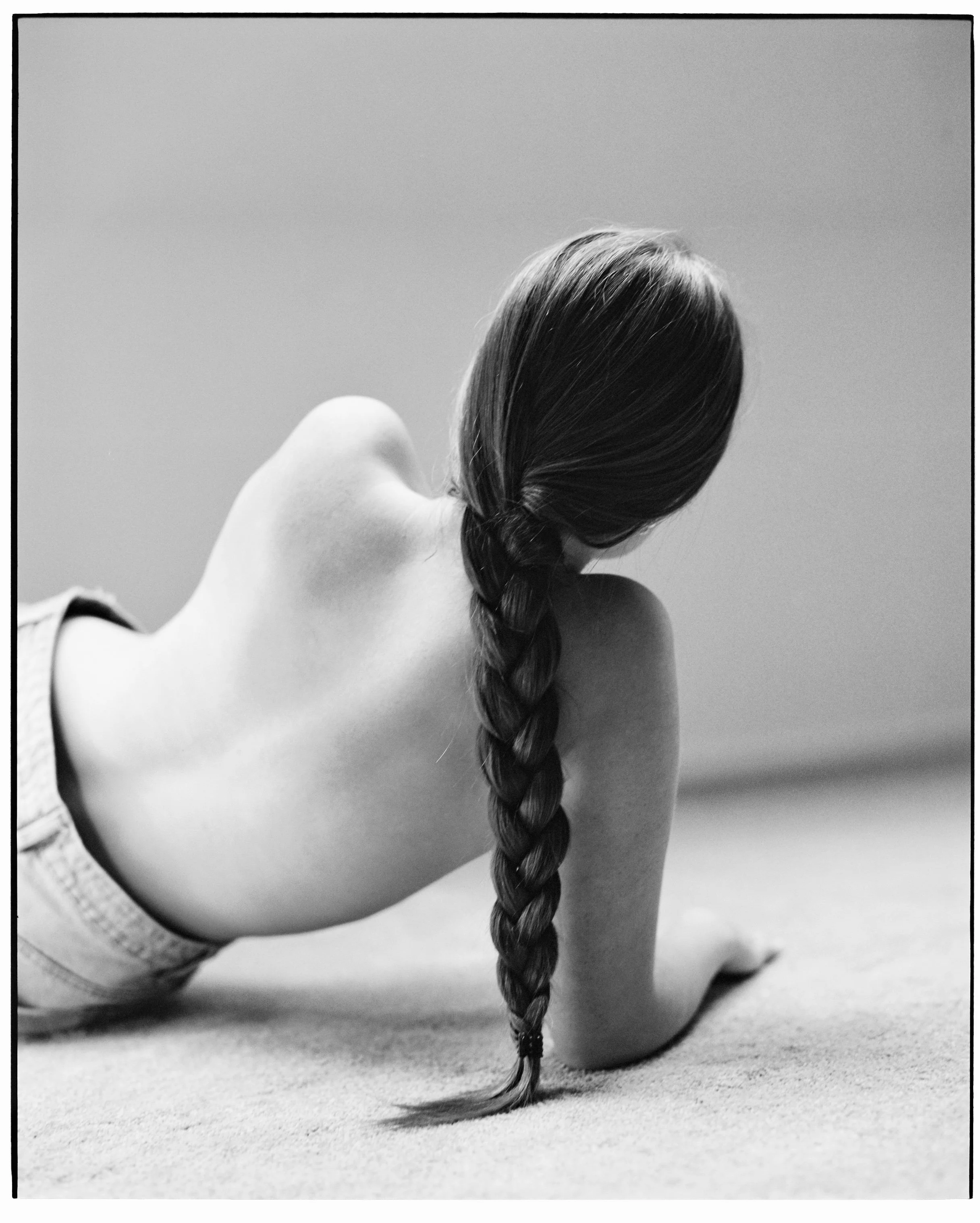 Black-and-white photo of a woman with long braided hair, lying on her stomach on a carpeted floor with her back to the camera, against a plain wall.