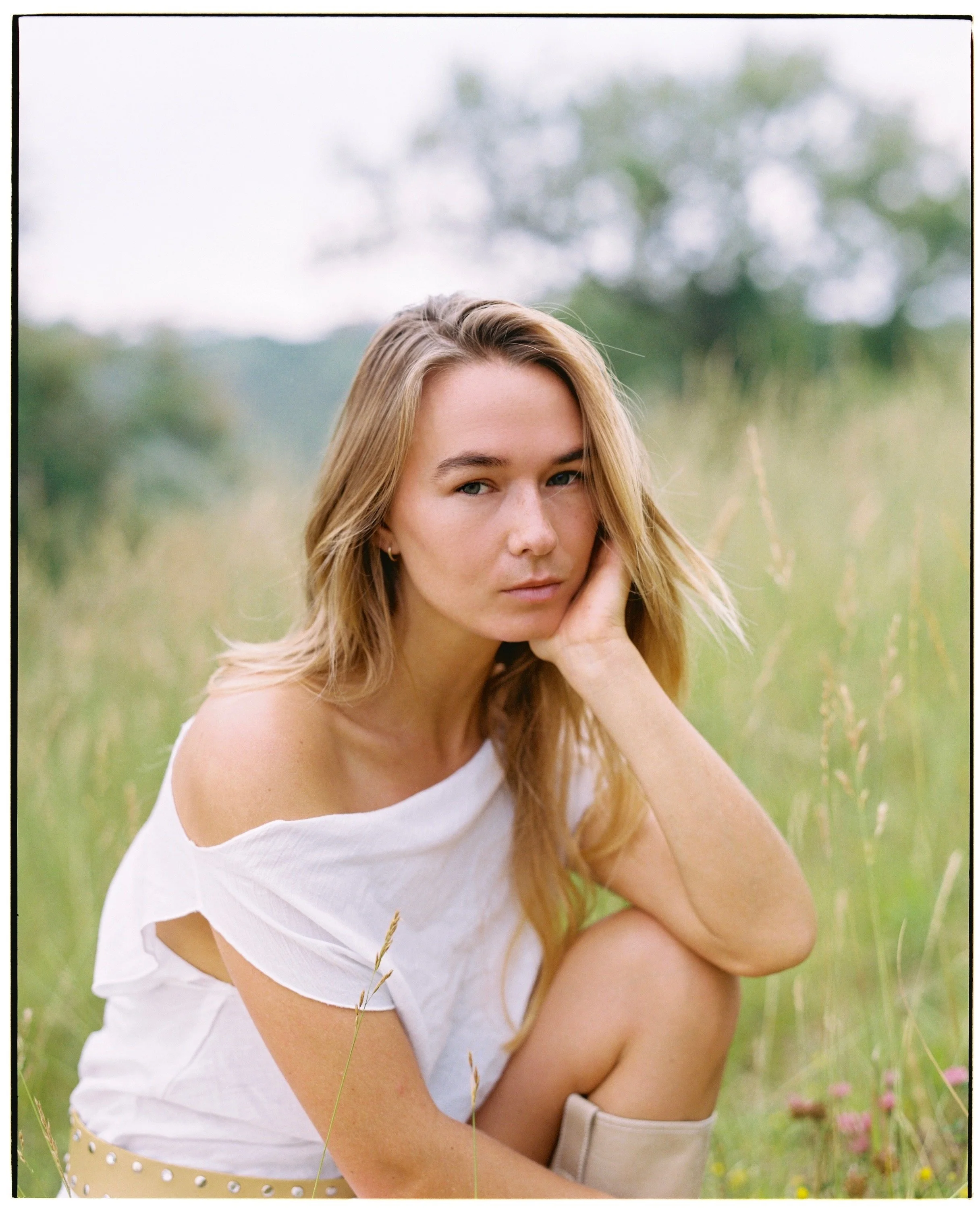 A young woman with blonde hair sitting in a grassy field, wearing a white off-shoulder top, with a contemplative expression, looking into the camera.