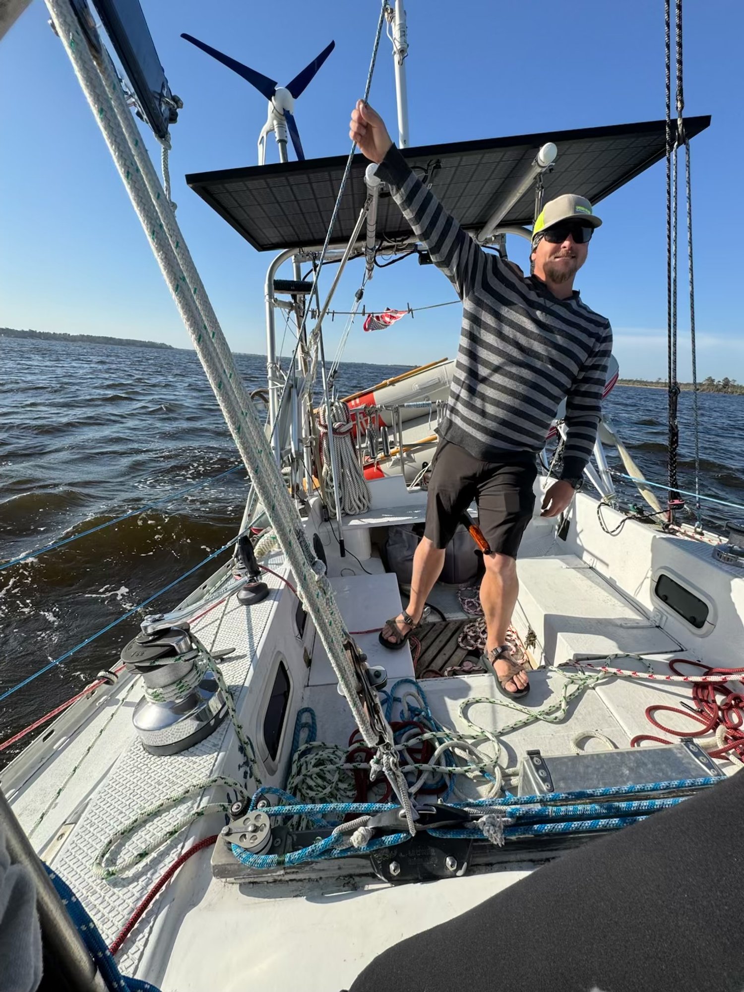 Captain helming a sailboat on deck, smiling, wearing sunglasses, a cap, a striped long-sleeve shirt, shorts, and sandals, holding a rope with his right hand, with water and sky in the background.