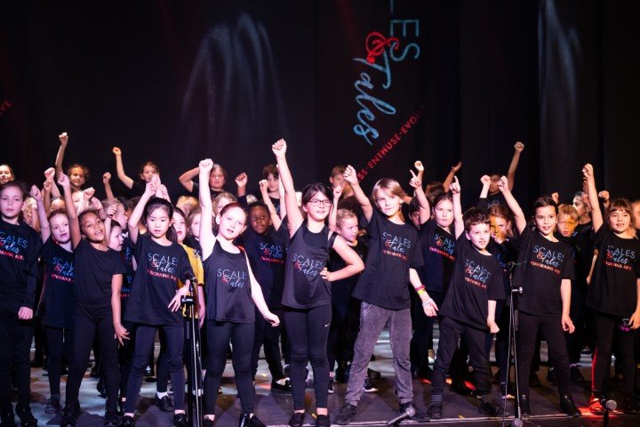 Children performing on stage at a dance recital, wearing matching black shirts and pants, with some raising fists as part of the dance.